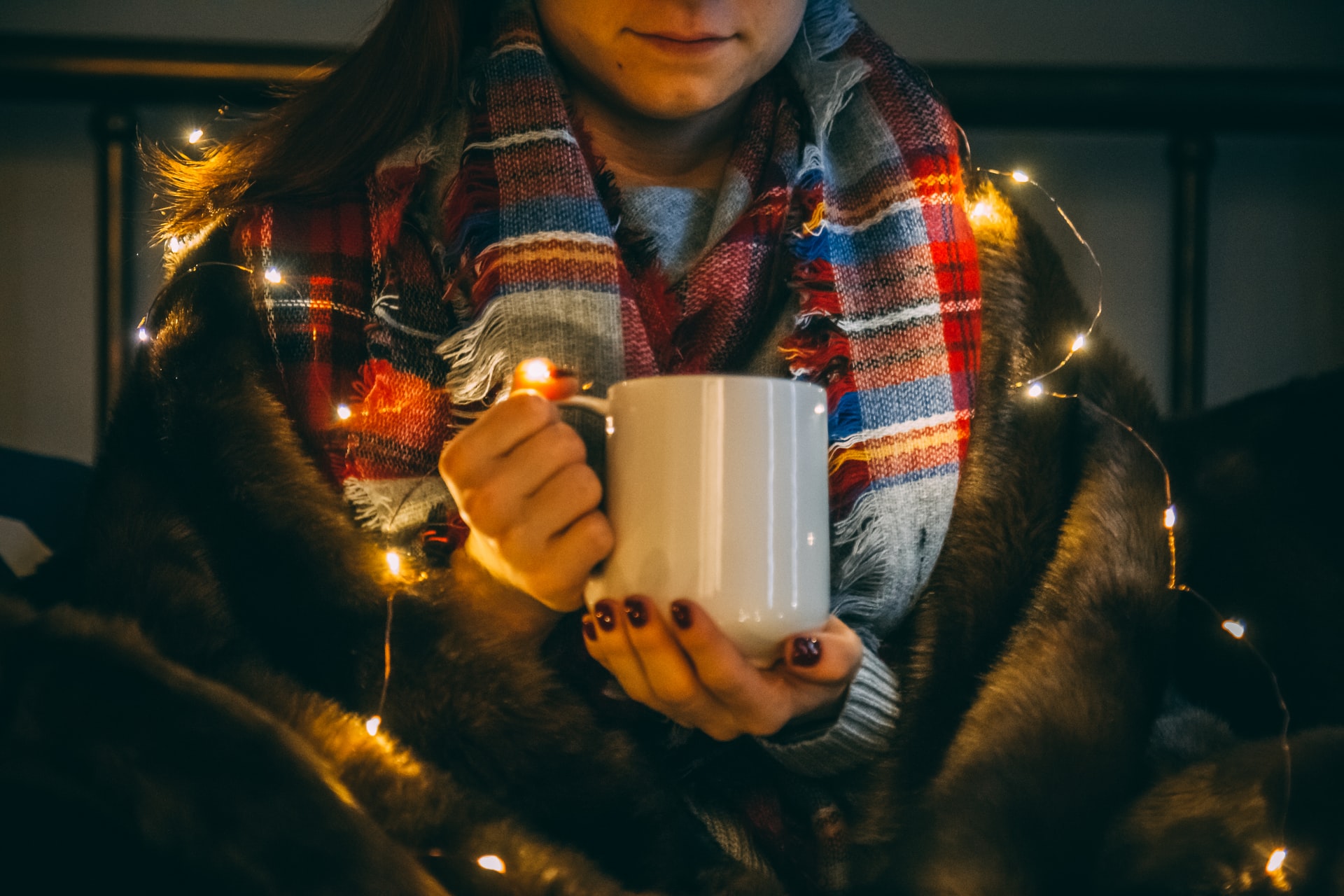 A girl in a festive scarf surrounded by Christmas lights holds a mug of hot rum punch.
