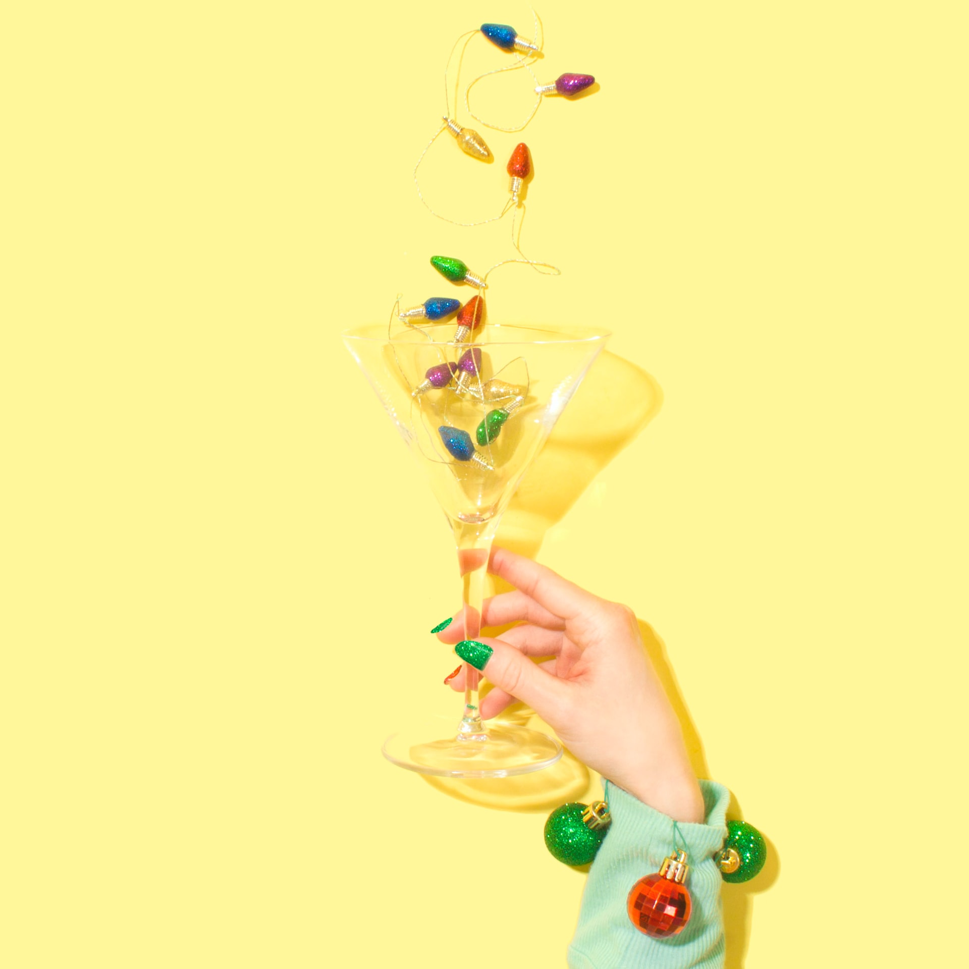 A hand with an ornament bracelet holds a martini glass filled with colorful Christmas lights against a bright yellow background.