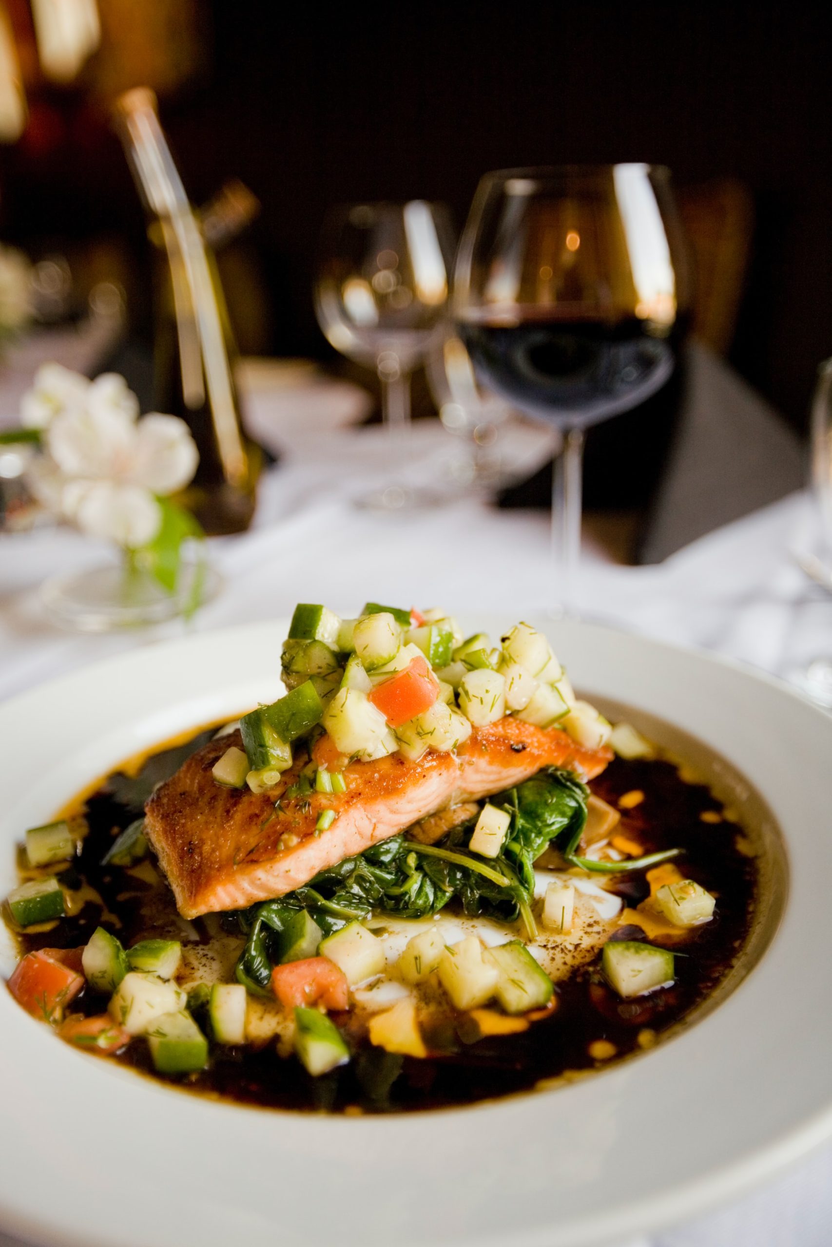 A salmon filet topped with tomato and cucumber relish on top of a bed of wilted greens sits on a table next to a glass of red wine.