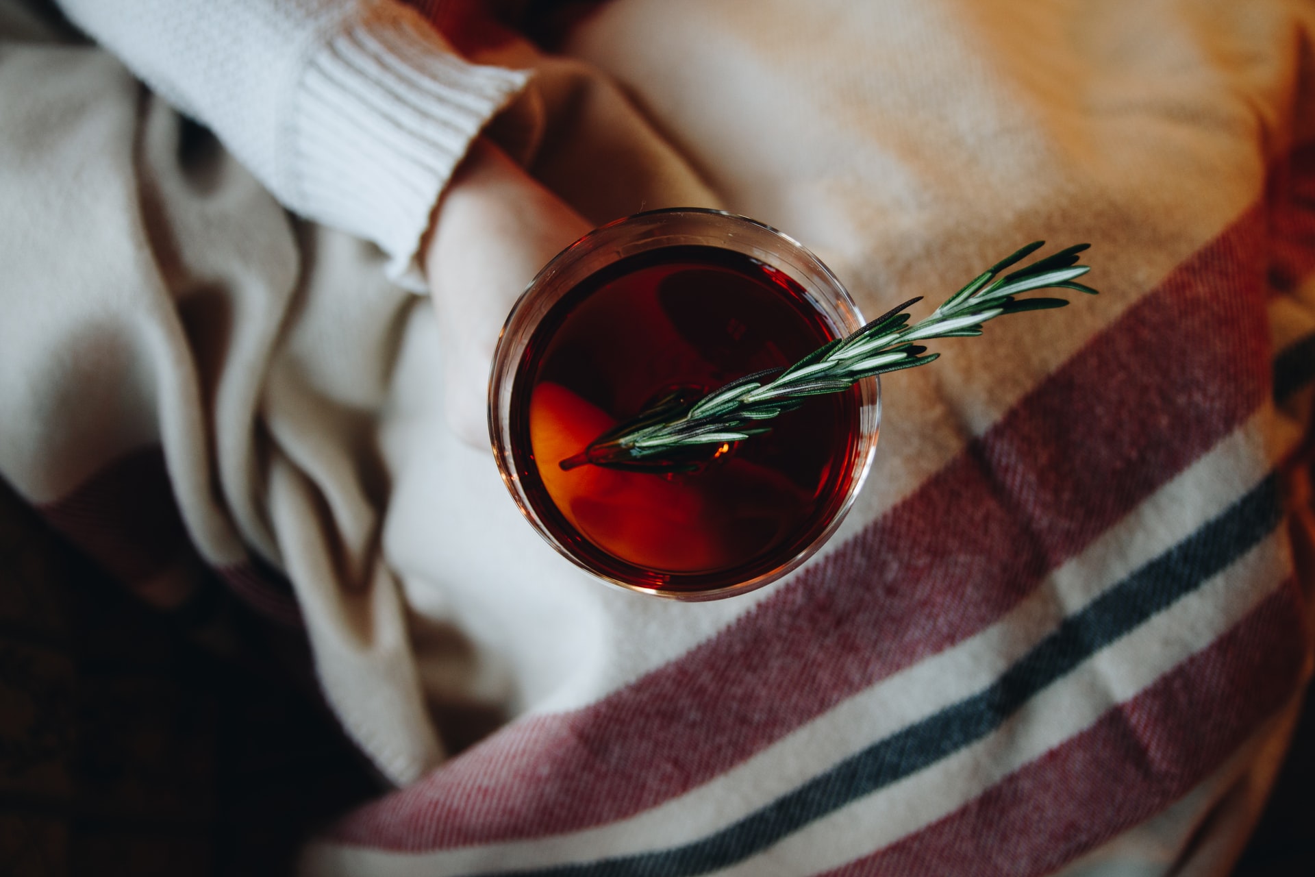 A person in a sweater under a blanket holds a glass of mulled wine with a sprig of rosemary.