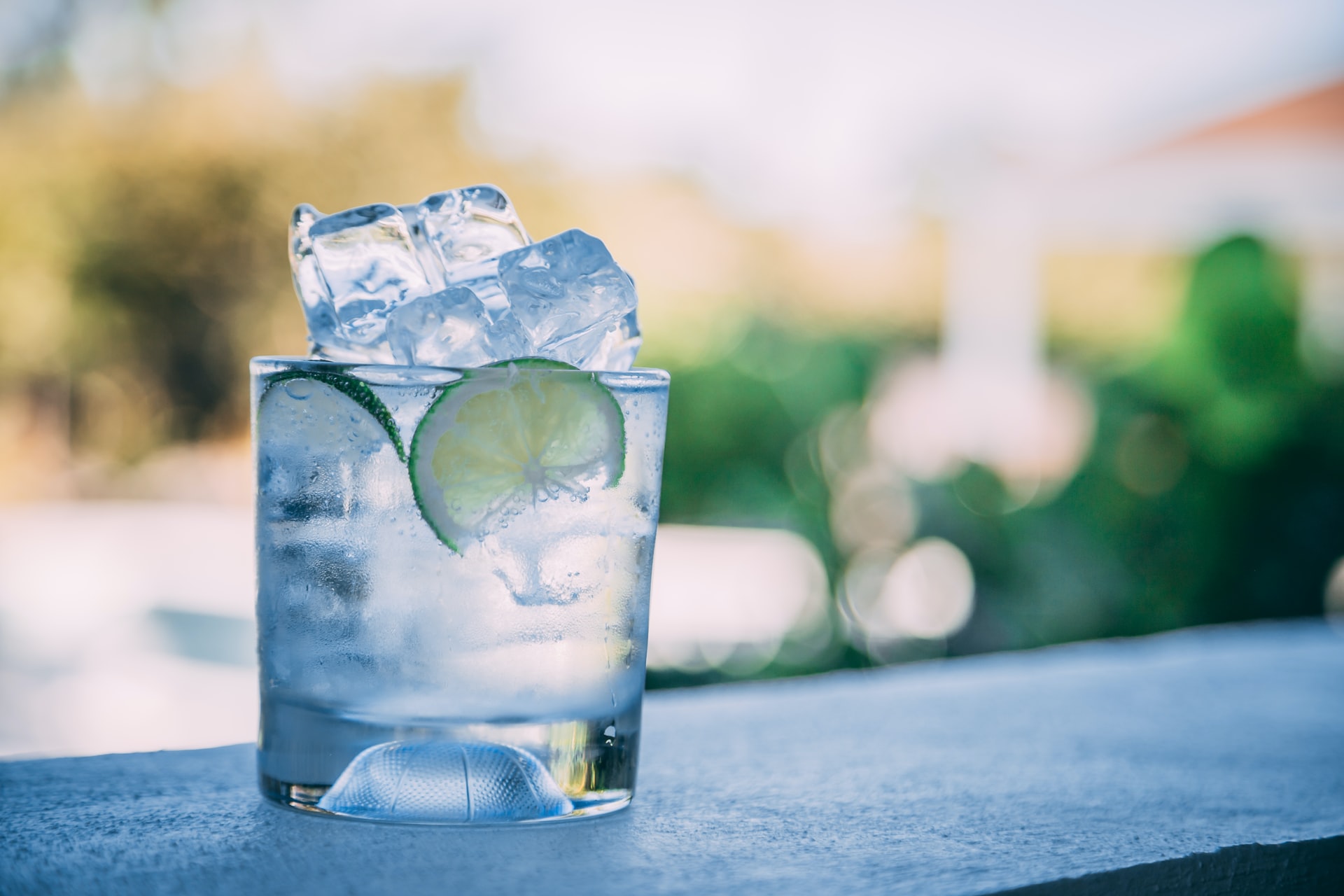 A rum and tonic in a glass with lime and ice sits on a ledge outside with a blurred background. 