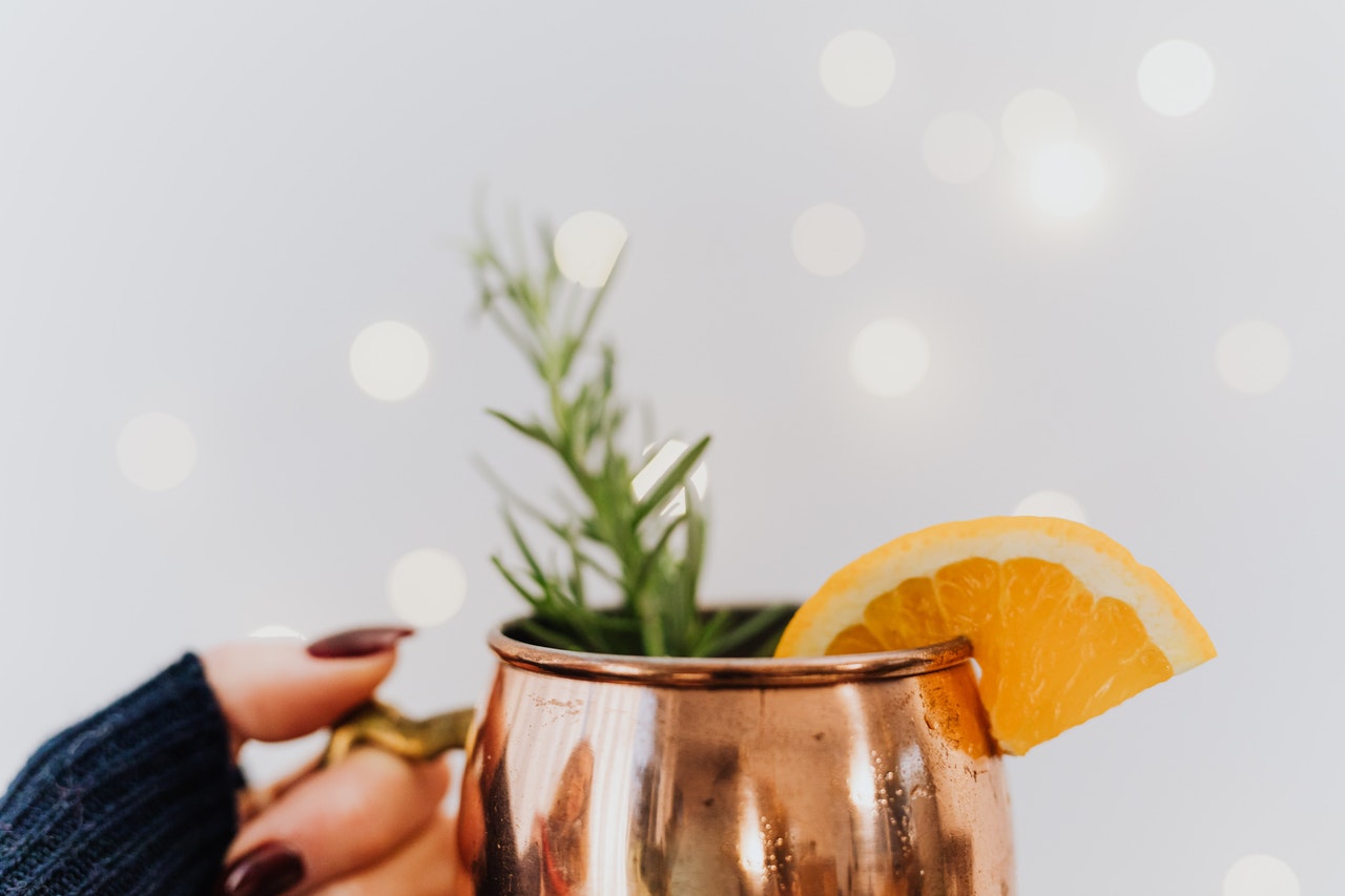 A hand holds a copper mug with hot mulled cider, rosemary, and an orange wedge.