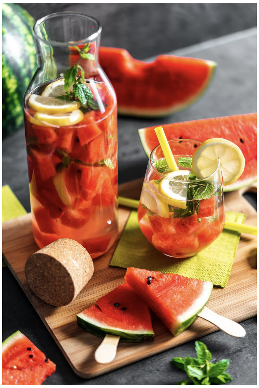 A pitcher and glass of watermelon punch garnished with watermelon slices, lemon wedges, and mint