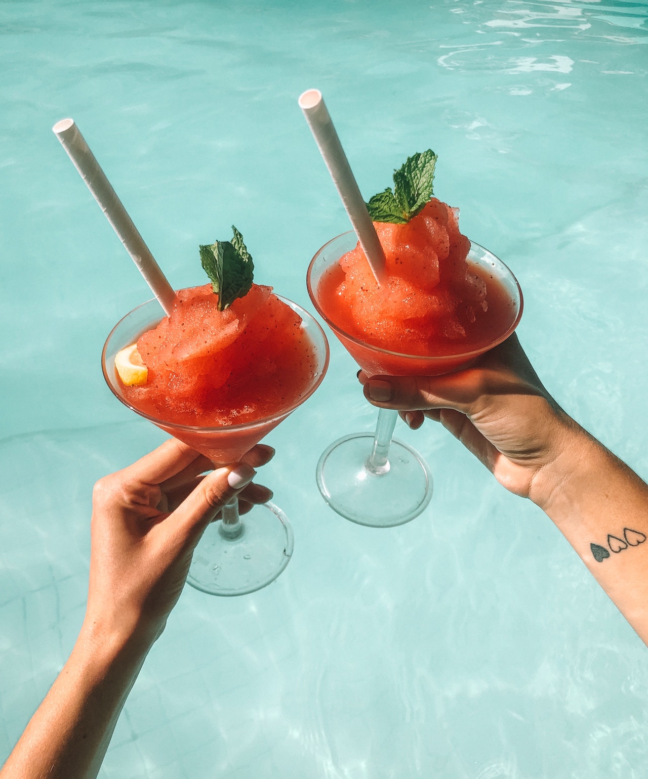 Two people cheers with frozen strawberry vodka slushies in front of a clear blue pool
