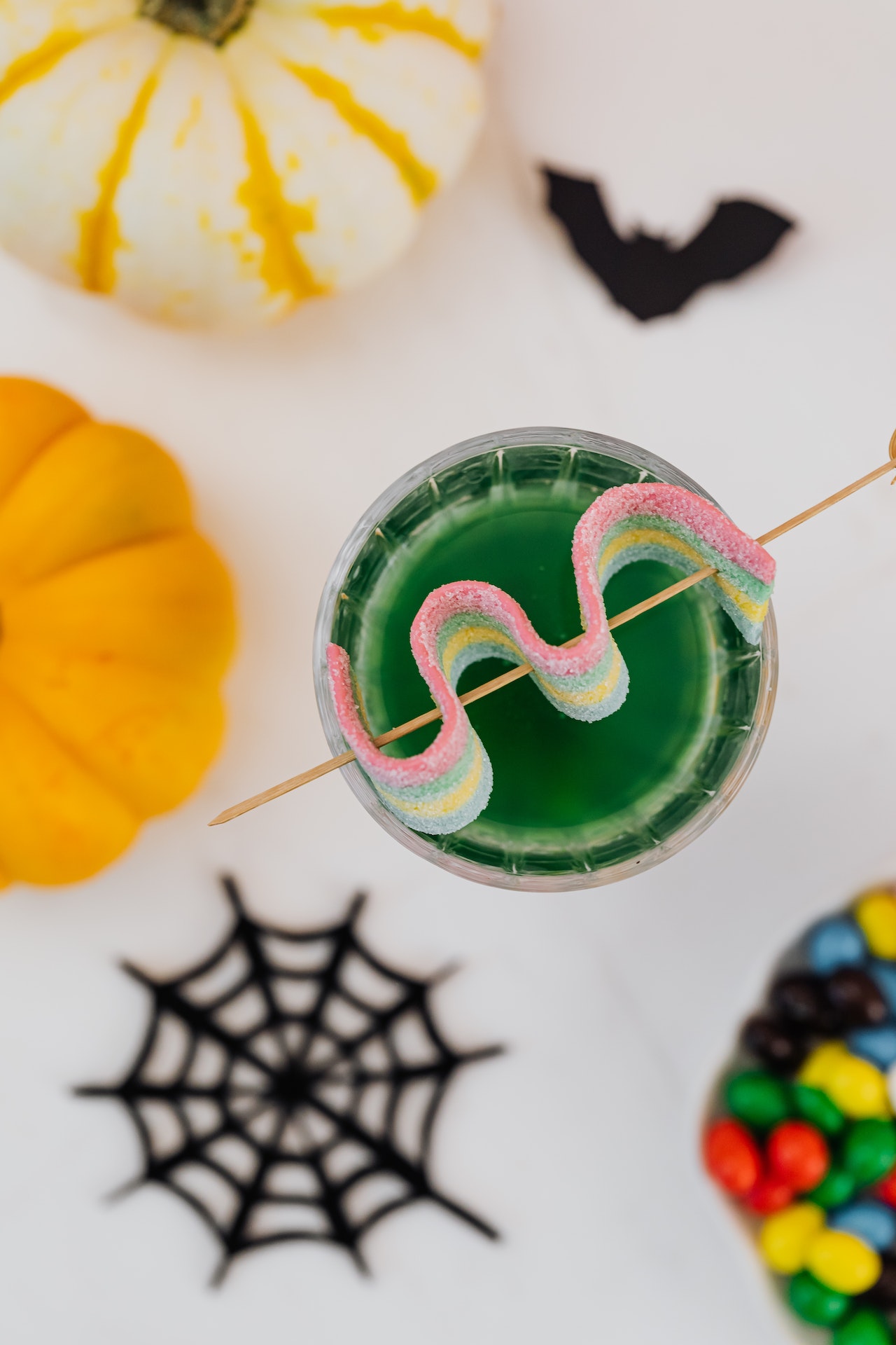 Aerial view of a bright green Halloween-themed cocktail with a gummy on top, surrounded by pumpkins, cobwebs, and bats.