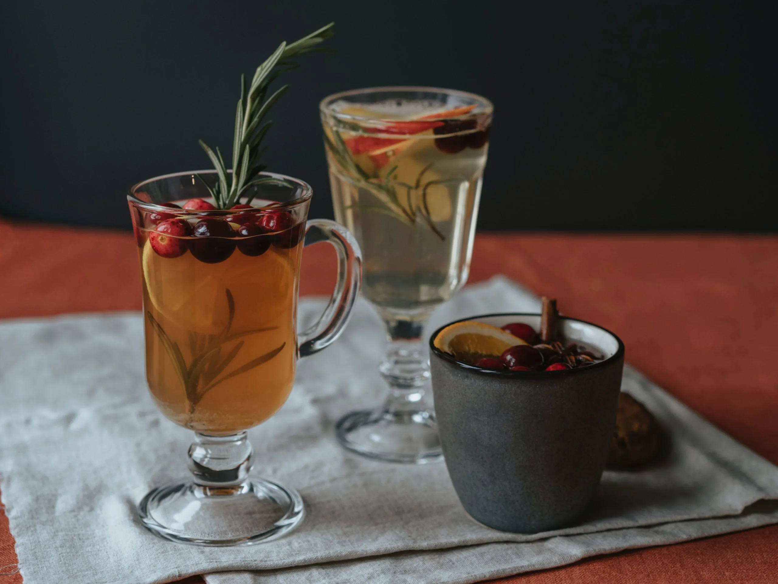 3 different hot toddies garnished with cranberries, rosemary, apple, orange, and cinnamon sticks.