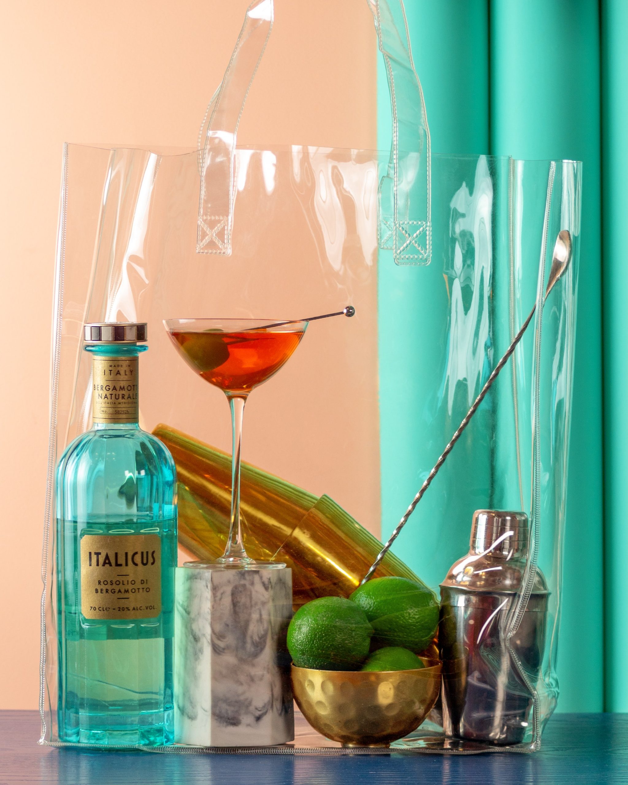 A bottle of Italicus, a martini, a bowl of limes, and cocktail shakers on a bar in a transparent bag.