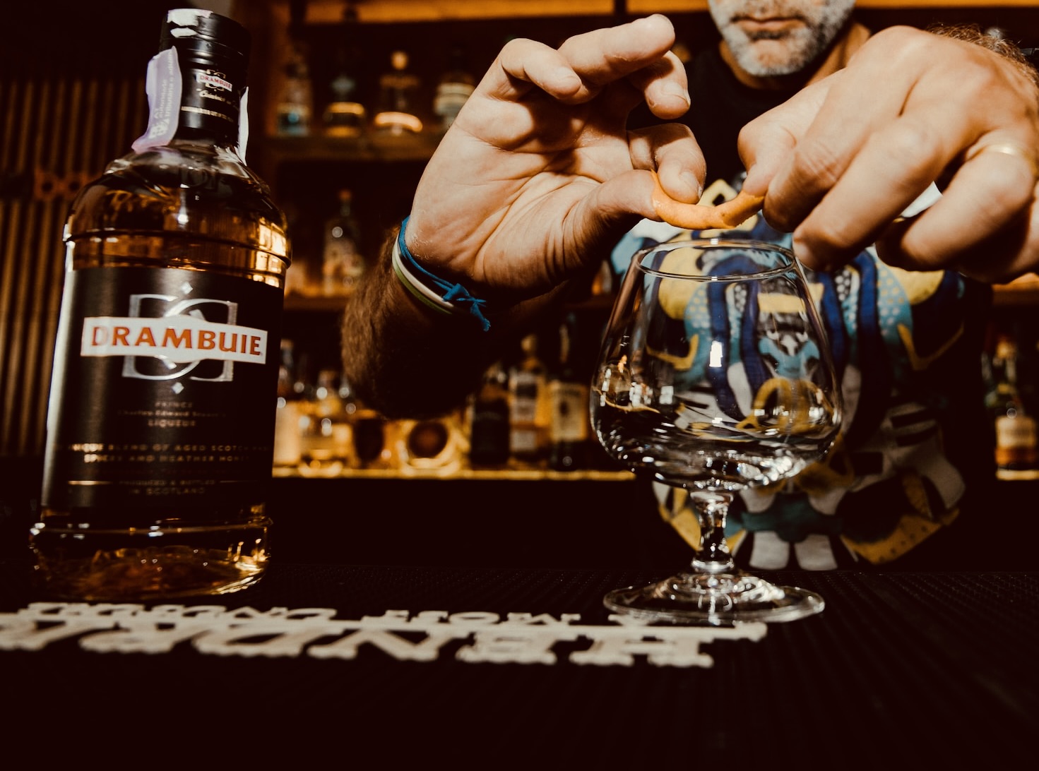 A bottle of Drambuie sits on a bar next to a man releasing oil from an orange peel into a glass 