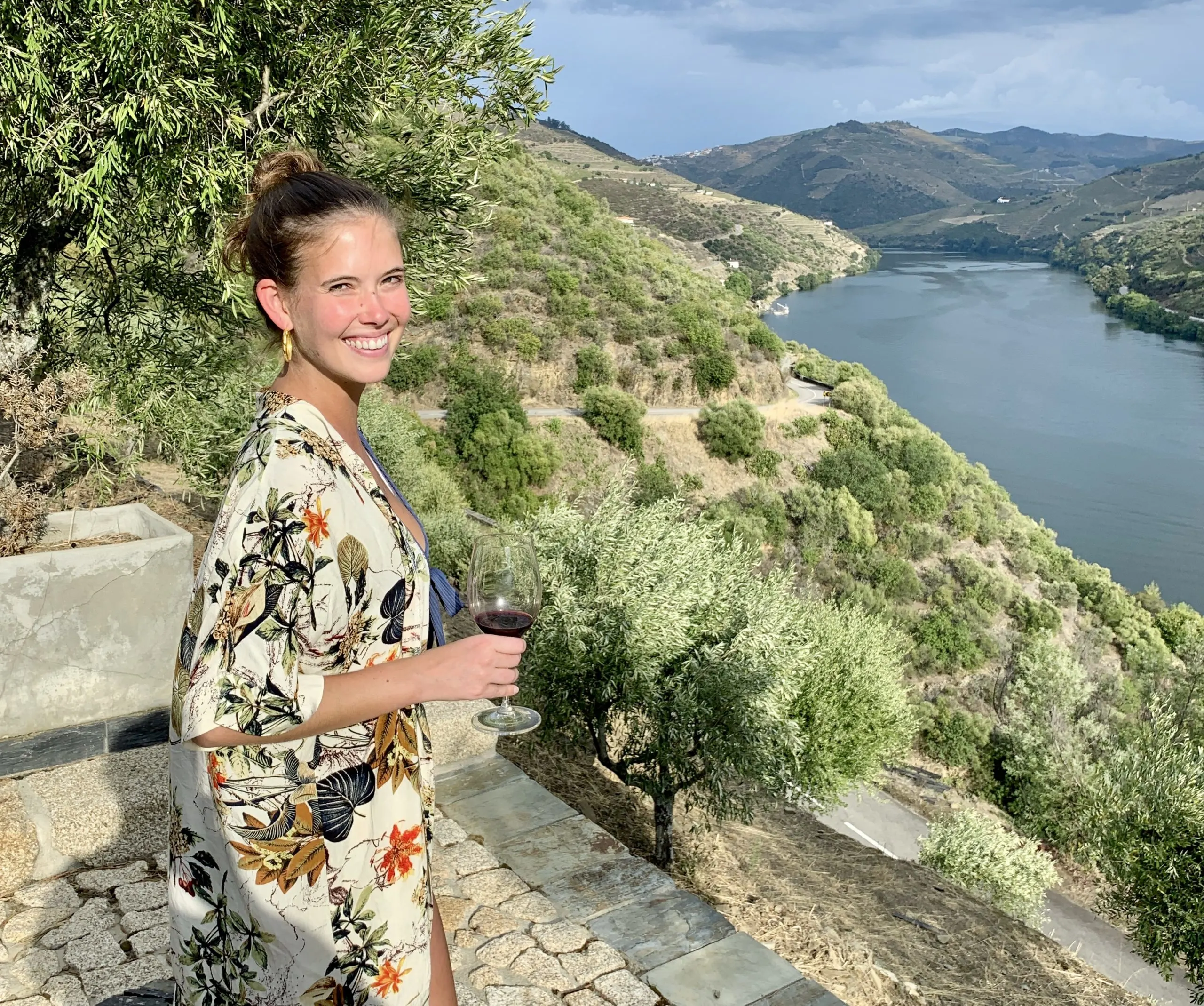 A smiling women holding a glass of red with the Douro river and wine region in the background.