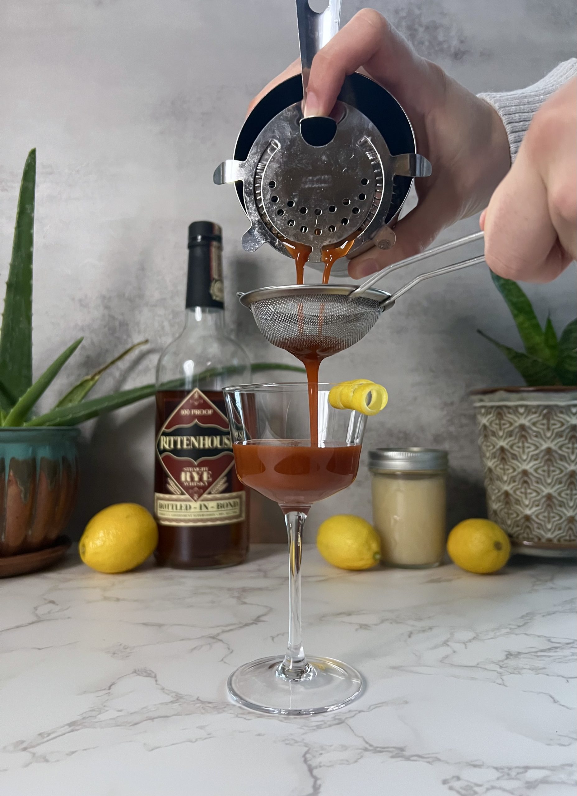A person double straining a Trinidad Sour cocktail from a Boston cocktail shaker through a Hawthorne strainer and fine mesh strainer with plants and lemons in the background.