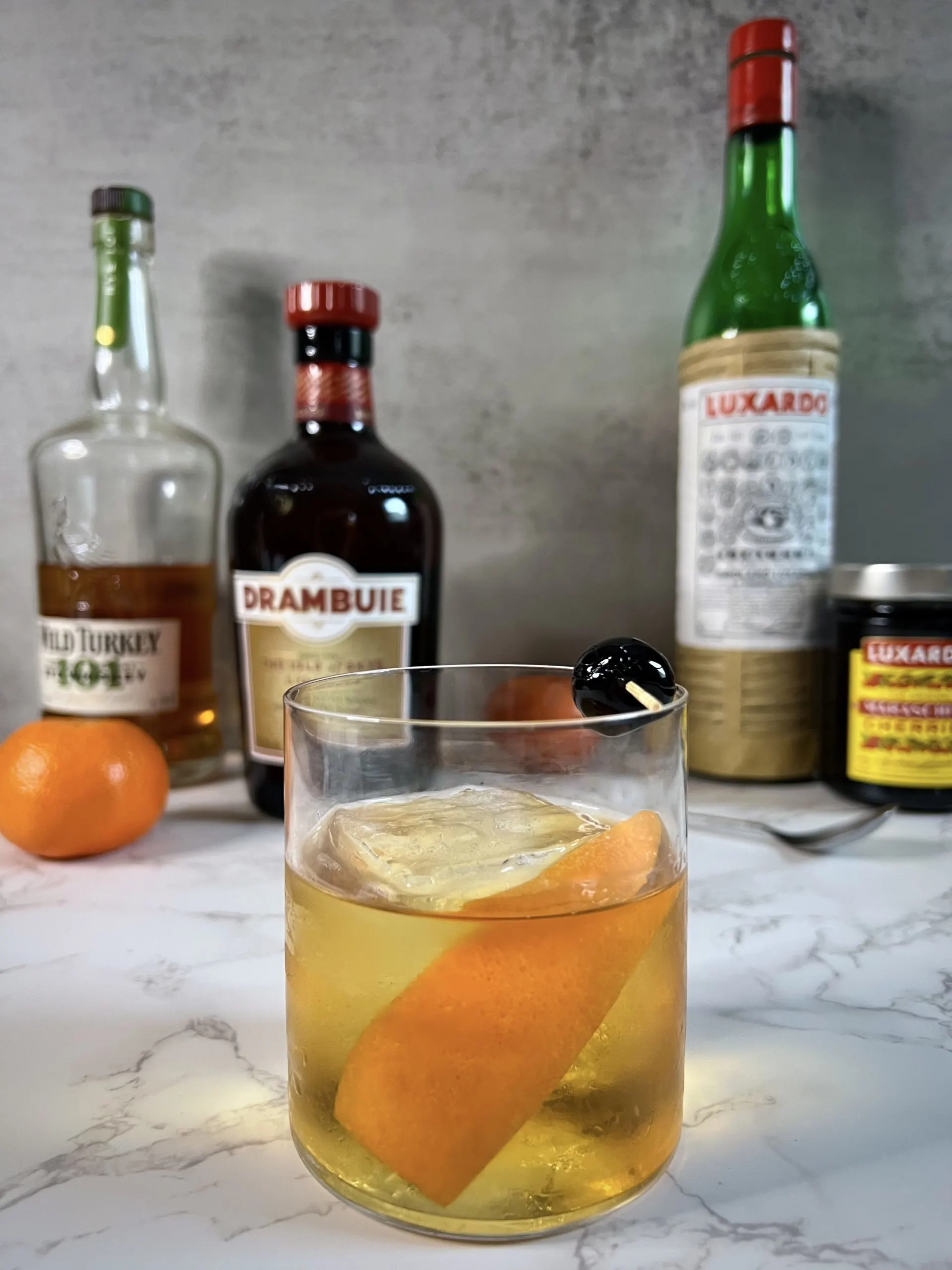 A Drambuie Old Fashioned with a Luxardo cherry in the foreground with liqueur bottles and oranges in the background.