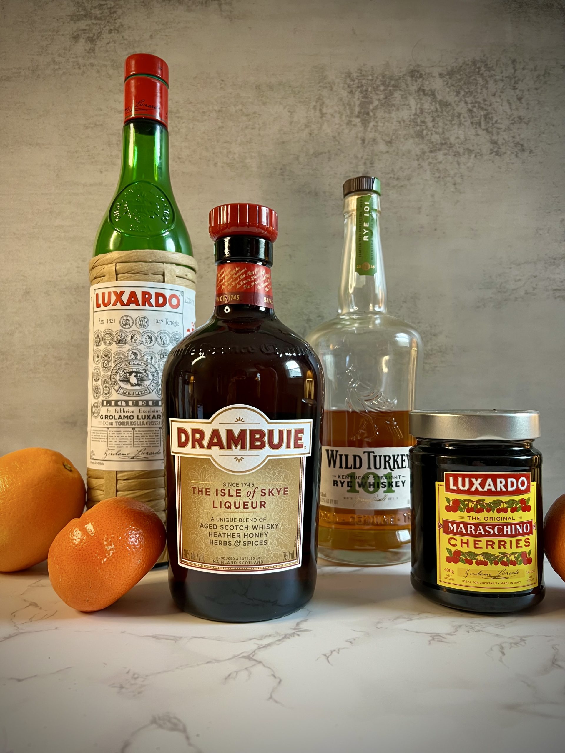A bottle of Luxardo Maraschino Originale, Drambuie, Wild Turkey Rye 101, and Luxardo cherries on a counter surrounded by oranges.