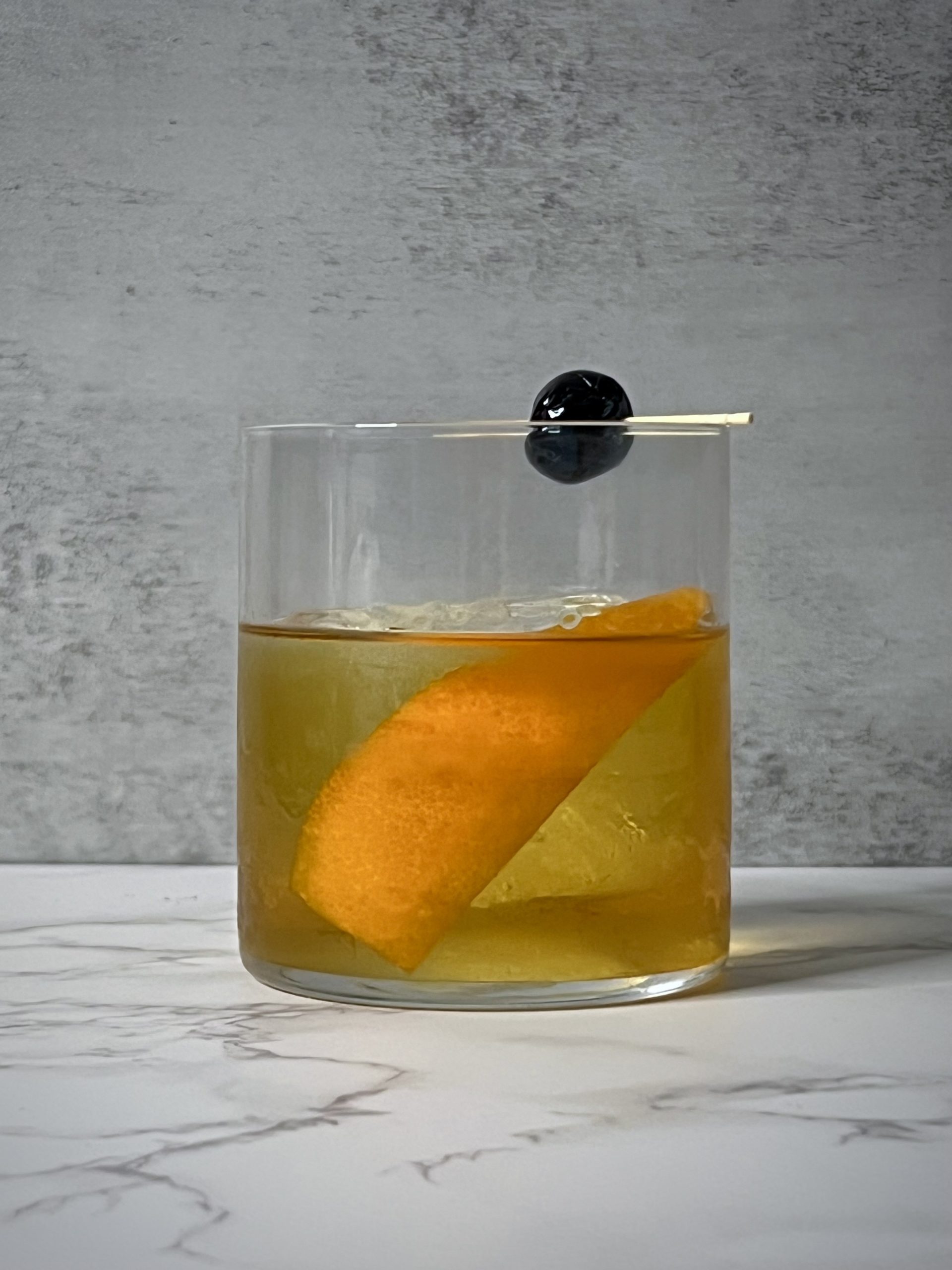 A straight on shot of a Drambuie Old Fashioned with an orange peel and Luxardo cherry on a toothpick against a grey background.