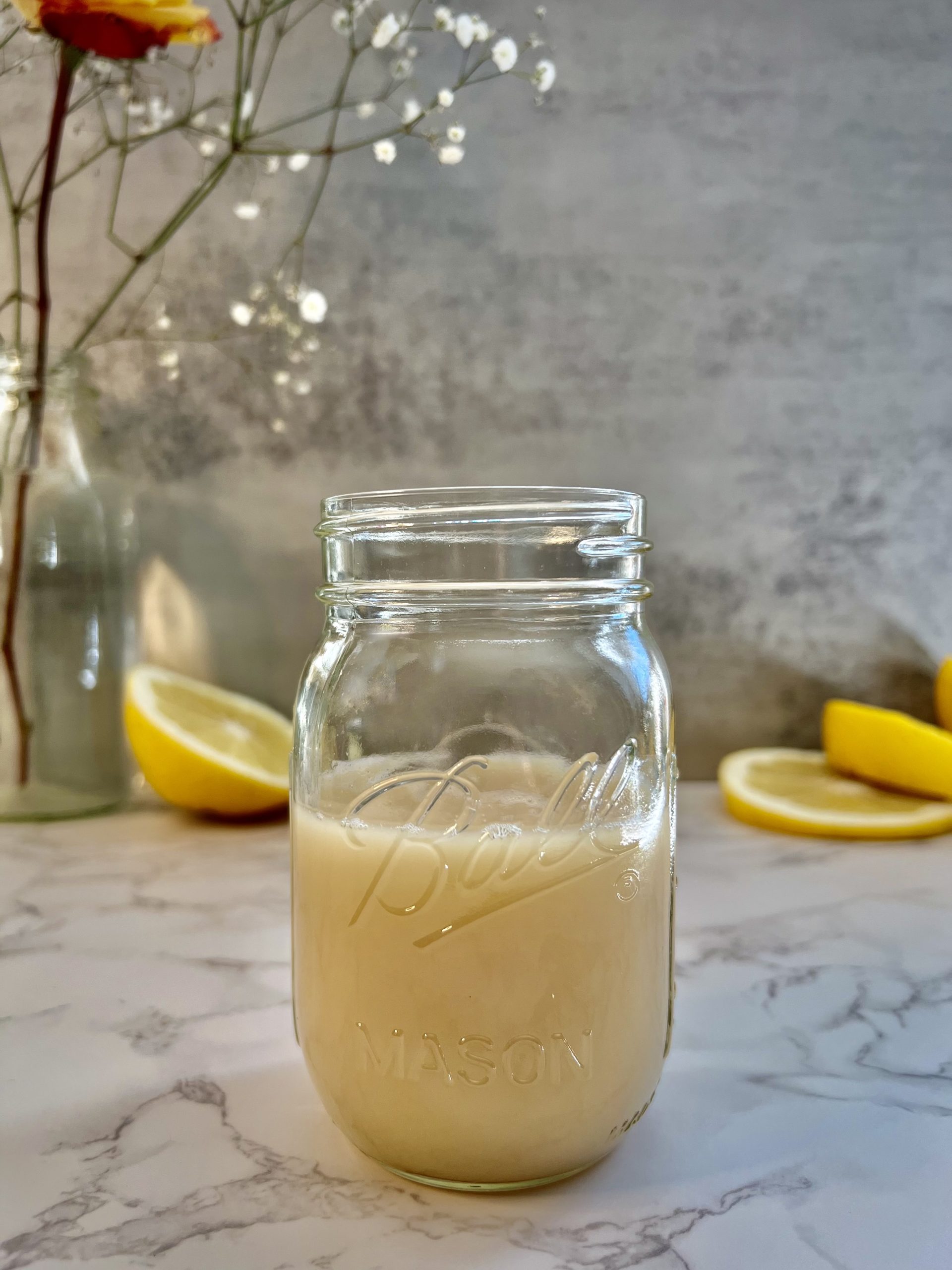 A mason jar filled with homemade orgeat on a counter with flowers and sliced white grapefruit in the background.