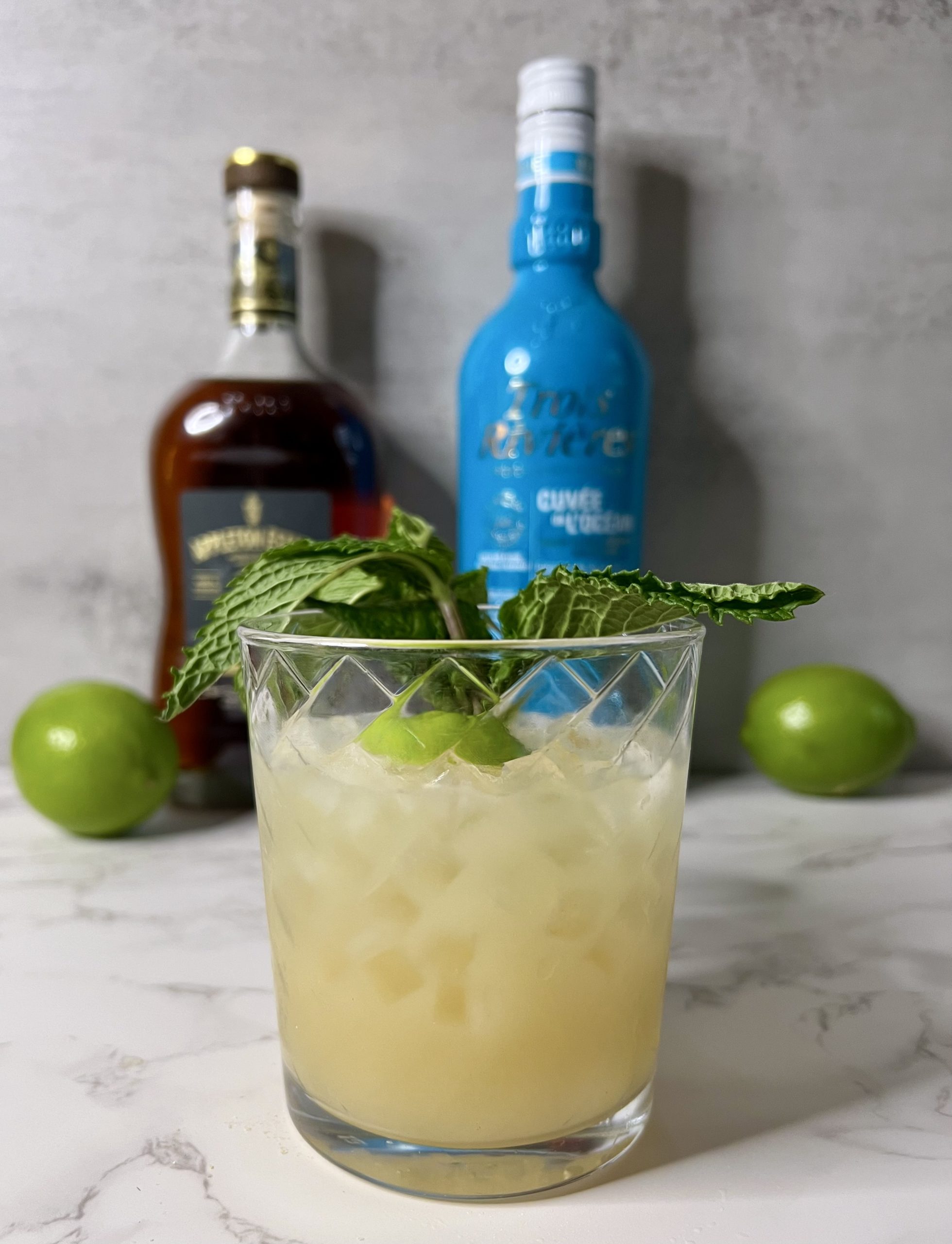 A Mai Tai garnished with a lime half and big sprig of mint in the foreground with two bottles of Caribbean rum and limes in the background.