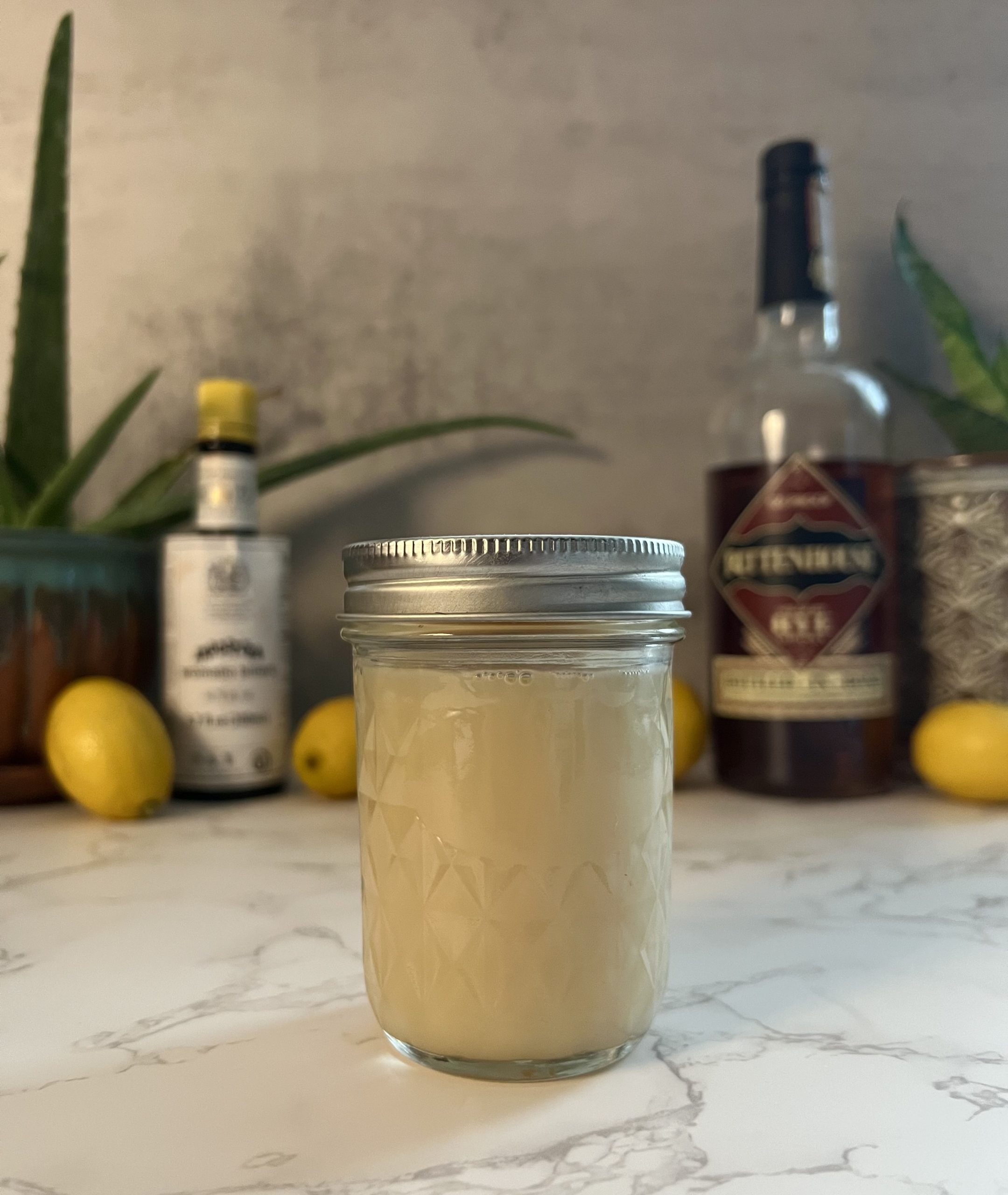 A mason jar filled with orgeat on a countertop with lemons, plants, and other ingredients blurred in the background.