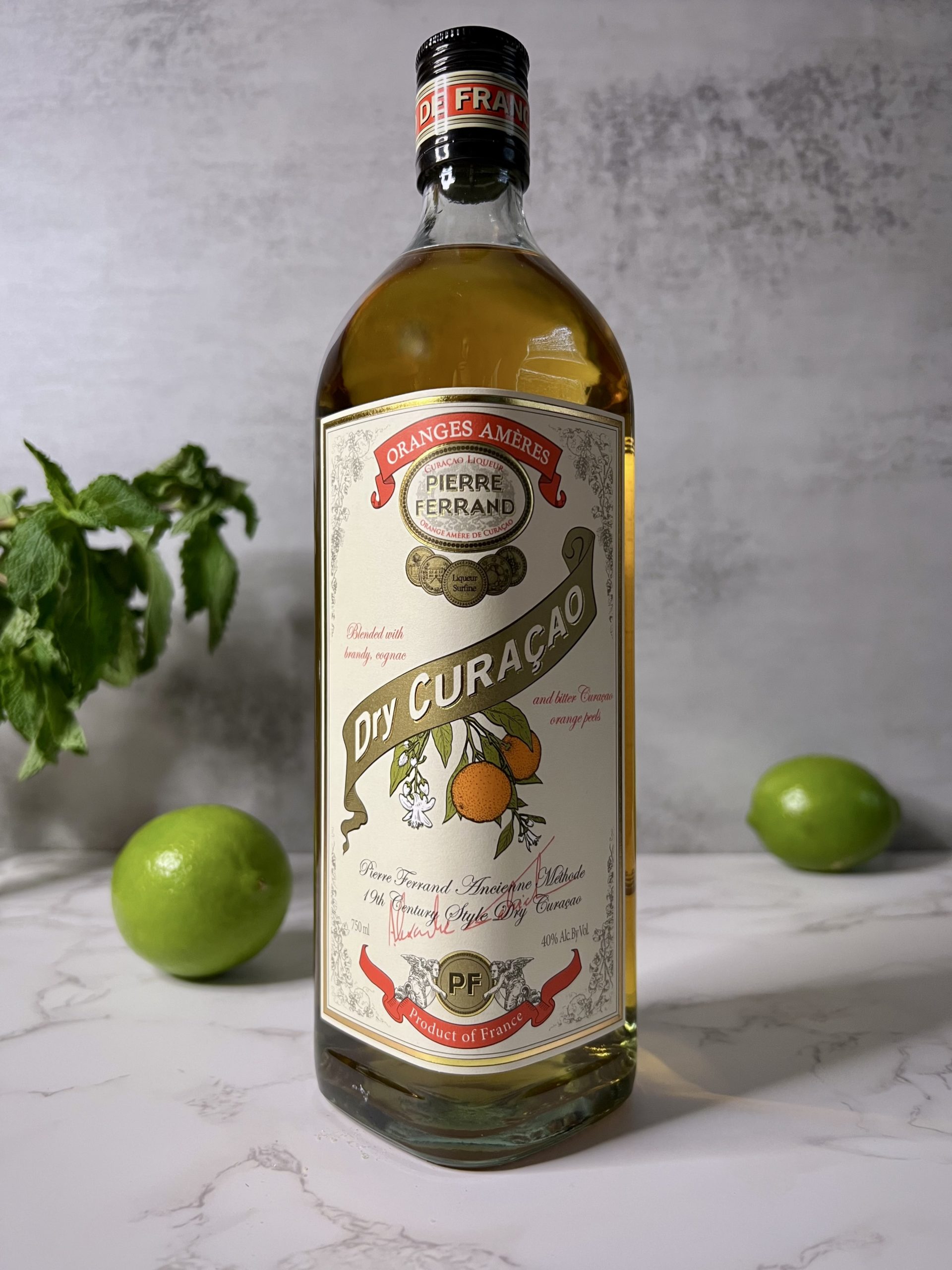 Close up of a bottle of Pierre Ferrand Dry Curacao with limes and mint in the background.