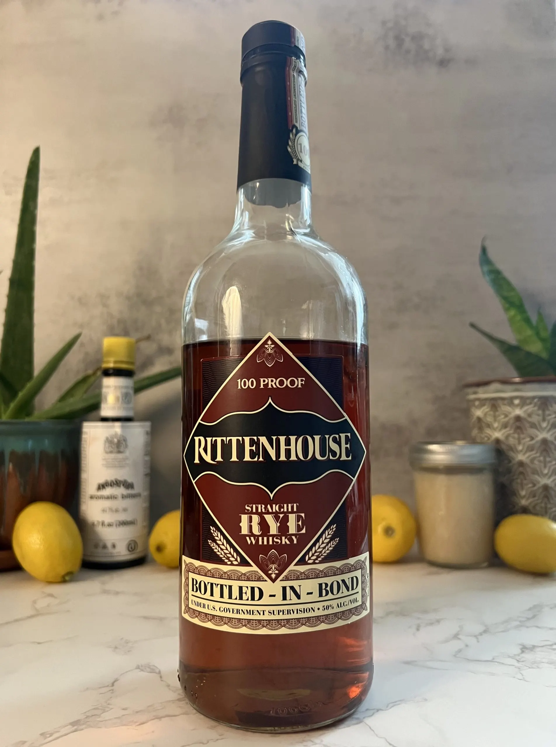 A close up shot of a bottle of Rittenhouse Straight Rye Whisky in the foreground with lemons, plants, and other ingredients in the background.