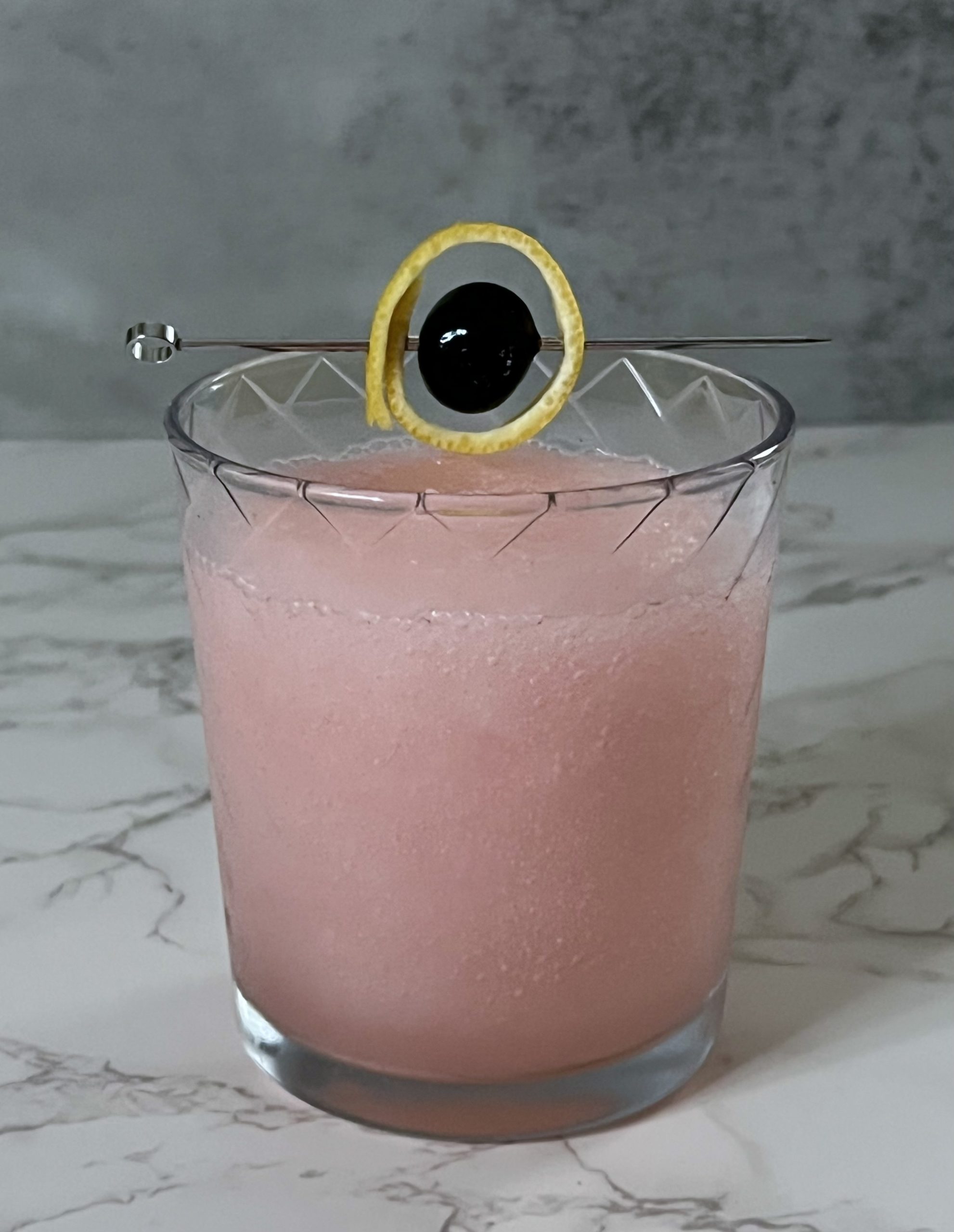 A frozen pink Saturn cocktail in a rocks glass on a countertop garnished with a lemon ring wrapped around a cherry on a cocktail pick to look like the planet Saturn.