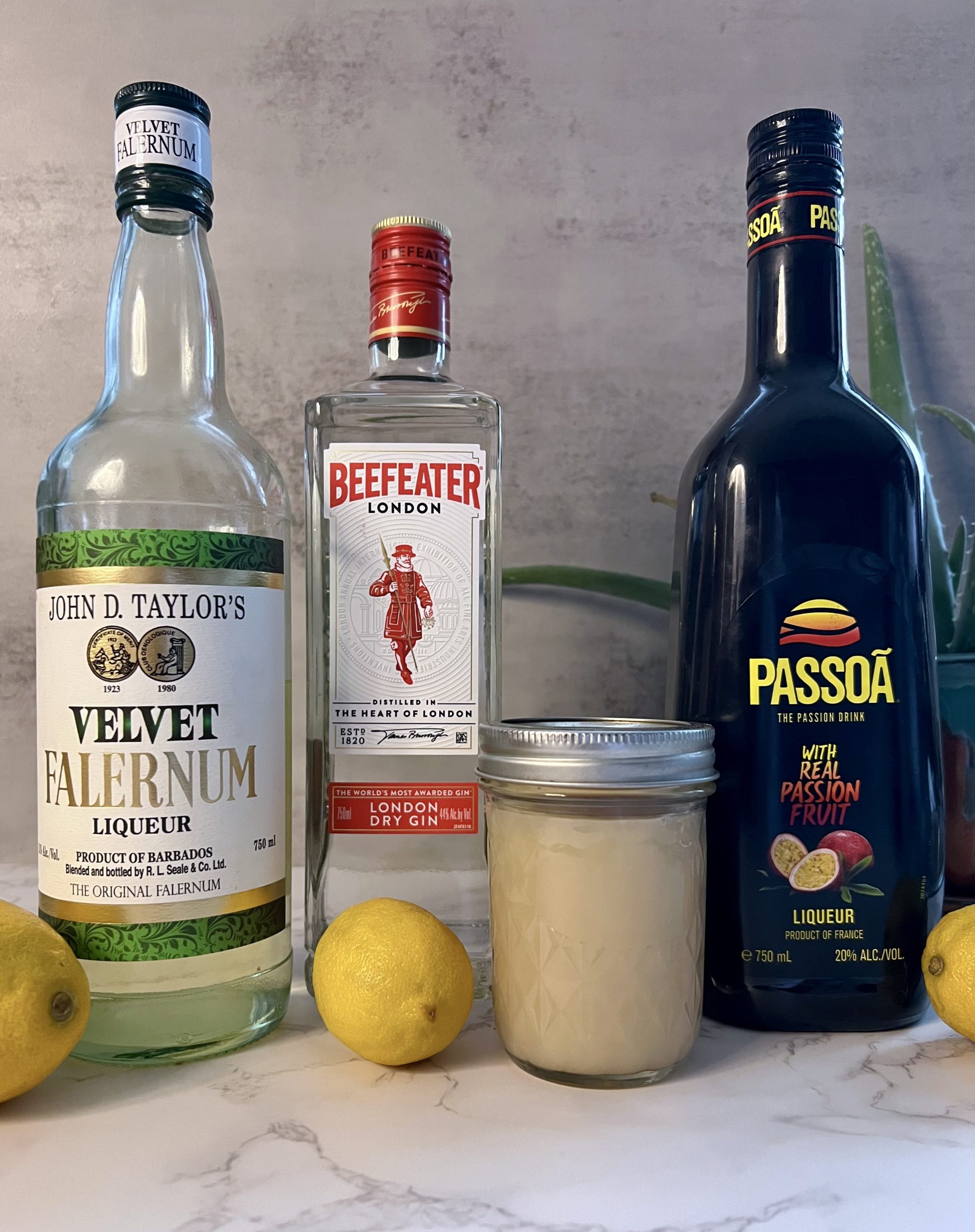 The ingredients of the Saturn cocktail, including Velvet Falernum, Beefeater Gin, orgeat syrup, Passoa Passion Fruit Liqueur, and lemons, lined up on a countertop.