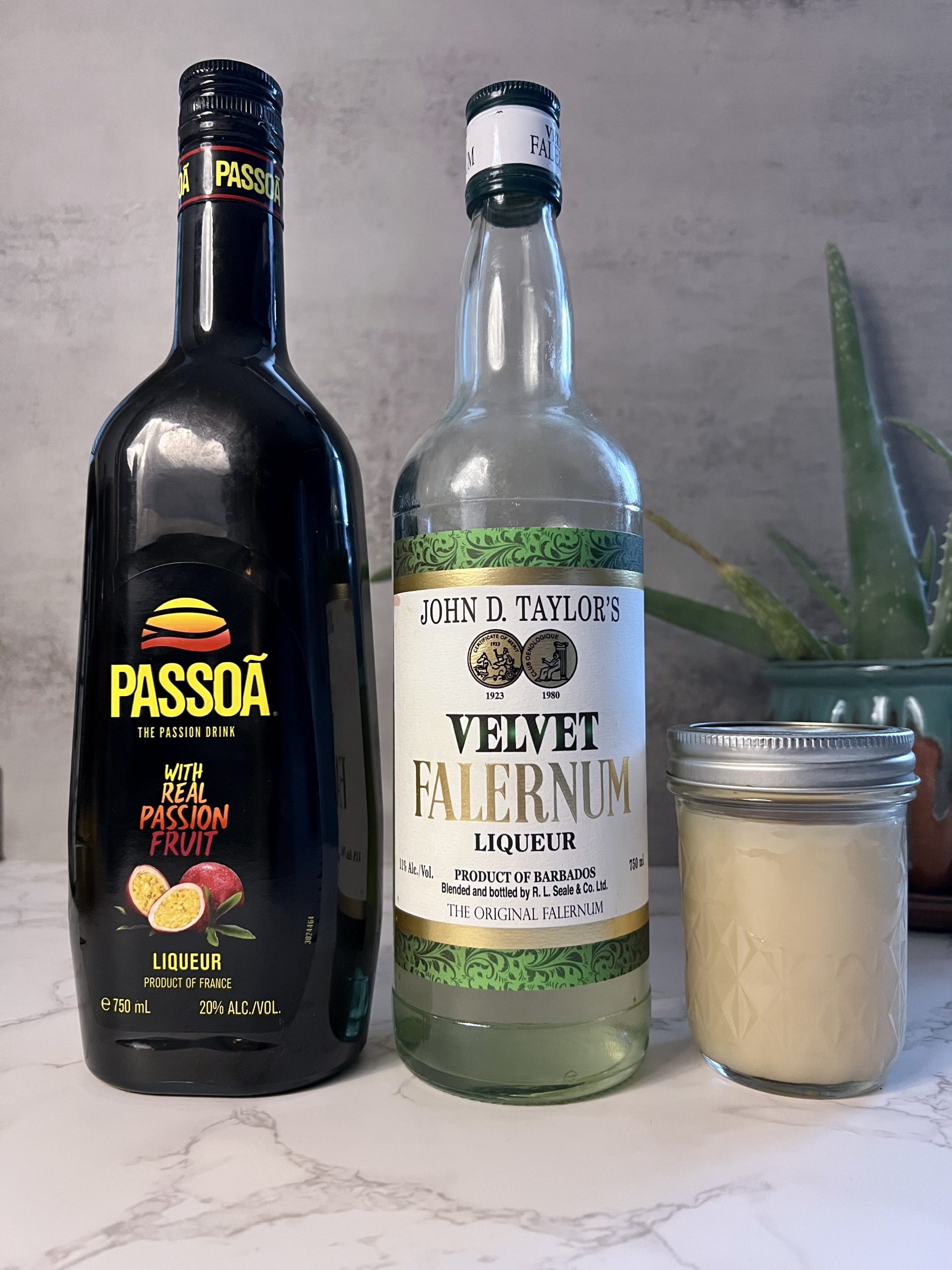 A bottle of Passoa Passion Fruit Liqueur, Velvet Falernum, and a mason jar of orgeat syrup lined up on a countertop with an aloe planet in the background.