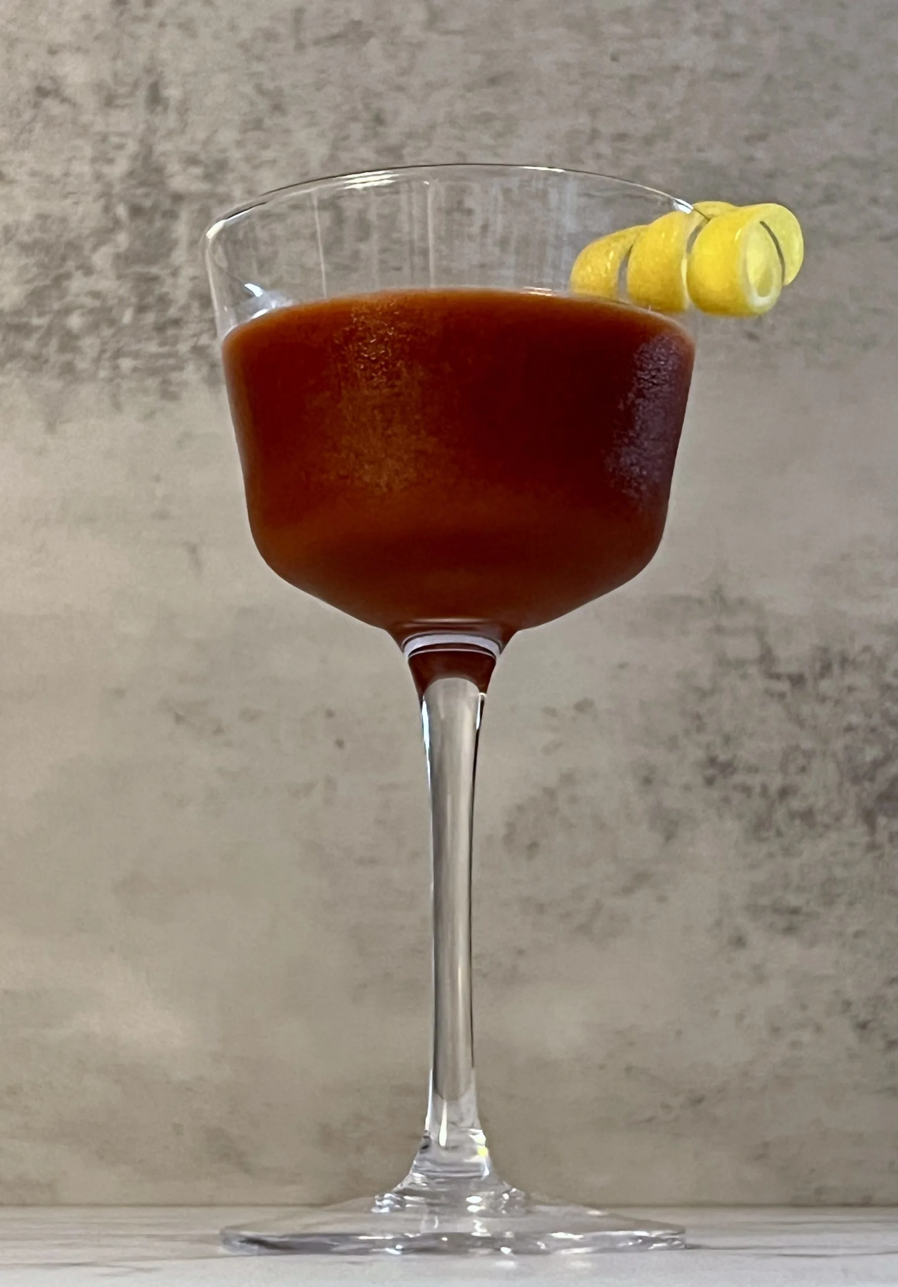 A dramatic close up shot of a vivid reddish-brown Trinidad Sour on a countertop garnished with a lemon twist.