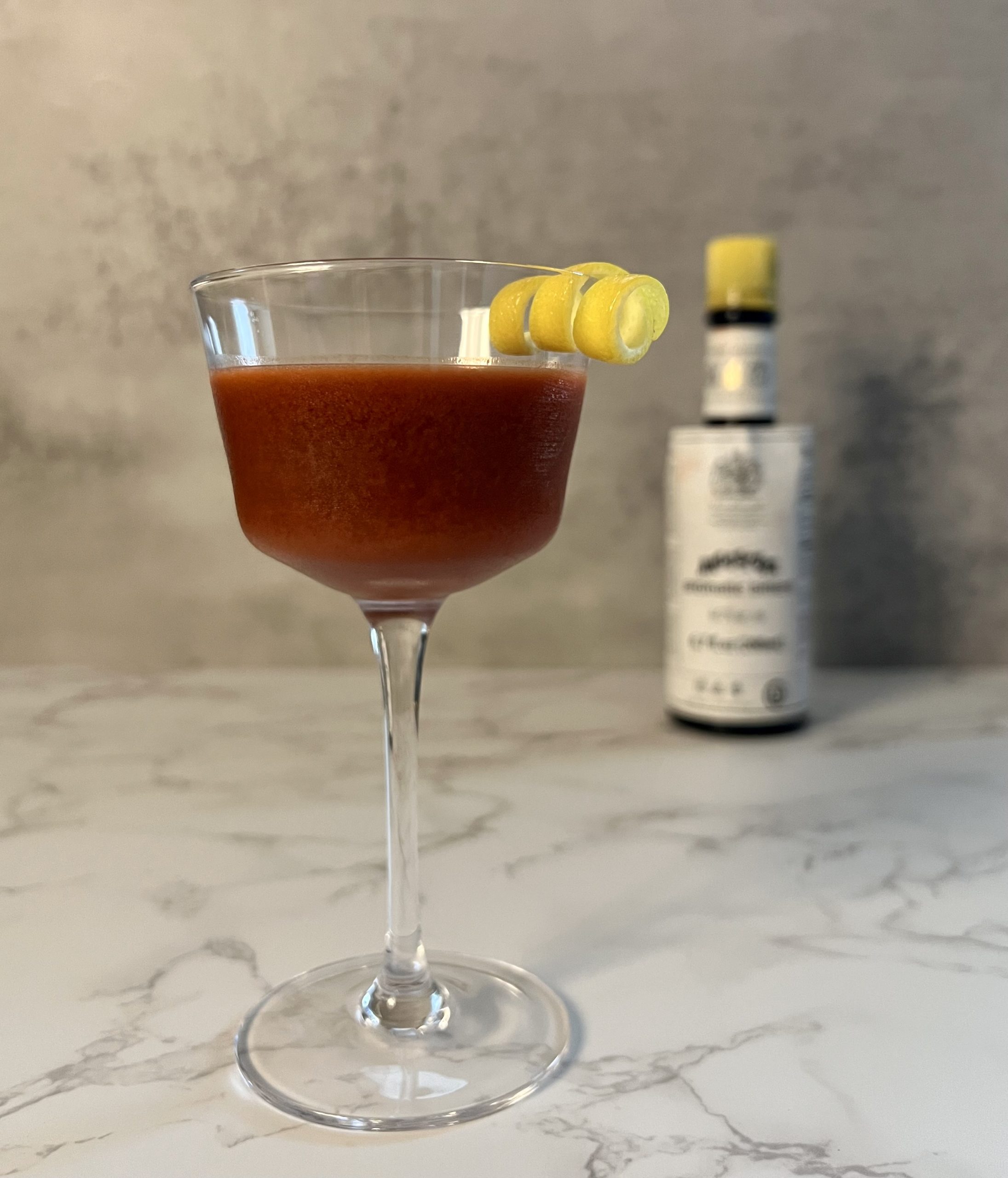 A Trinidad Sour garnished with a lemon twist in the foreground with a bottle of Angostura bitters behind it blurred in the background.
