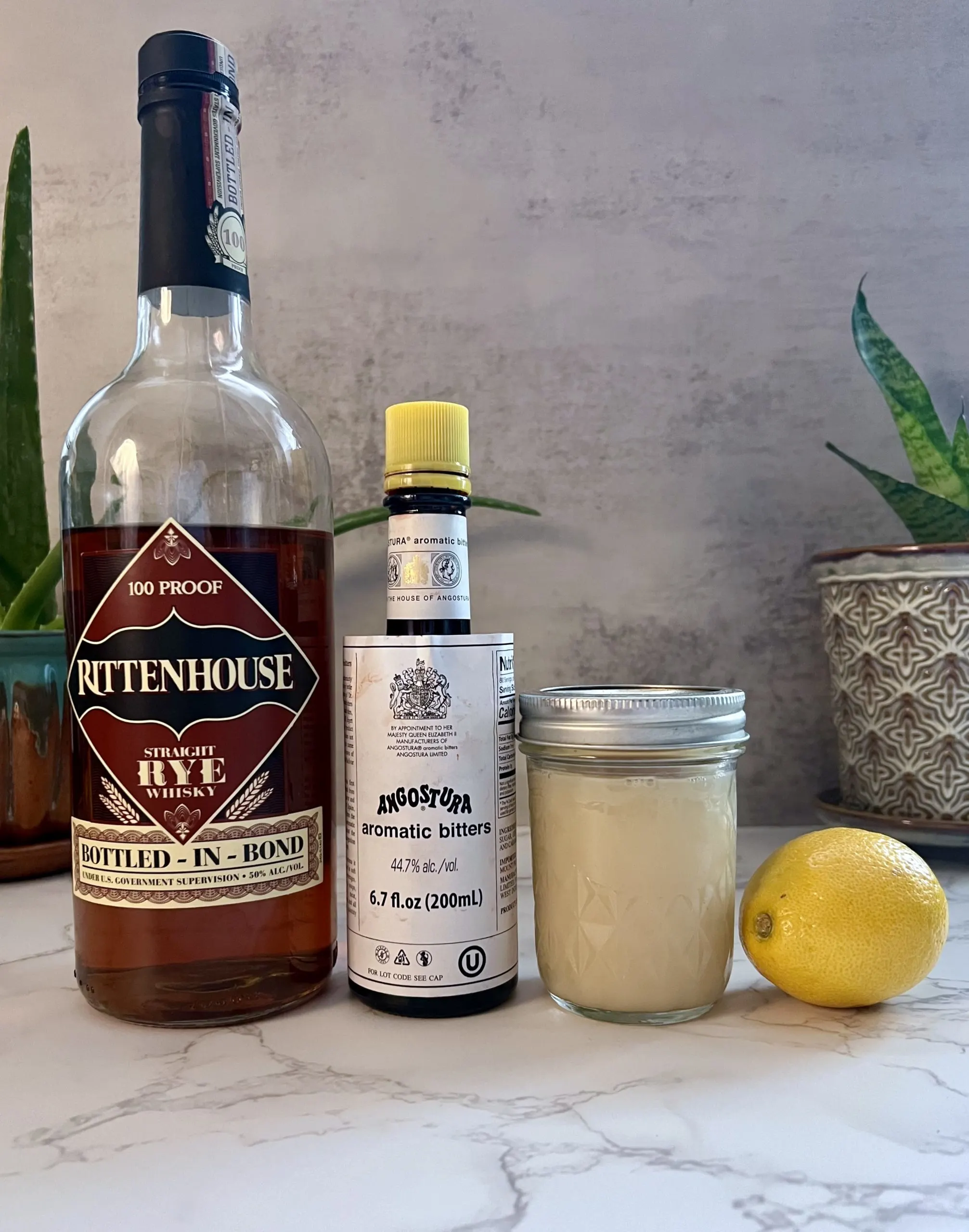 The ingredients of a Trinidad Sour, including a bottle of Rittenhouse Straight Rye Whisky, Angostura bitters, orgeat, and a lemon, all lined up on a countertop.