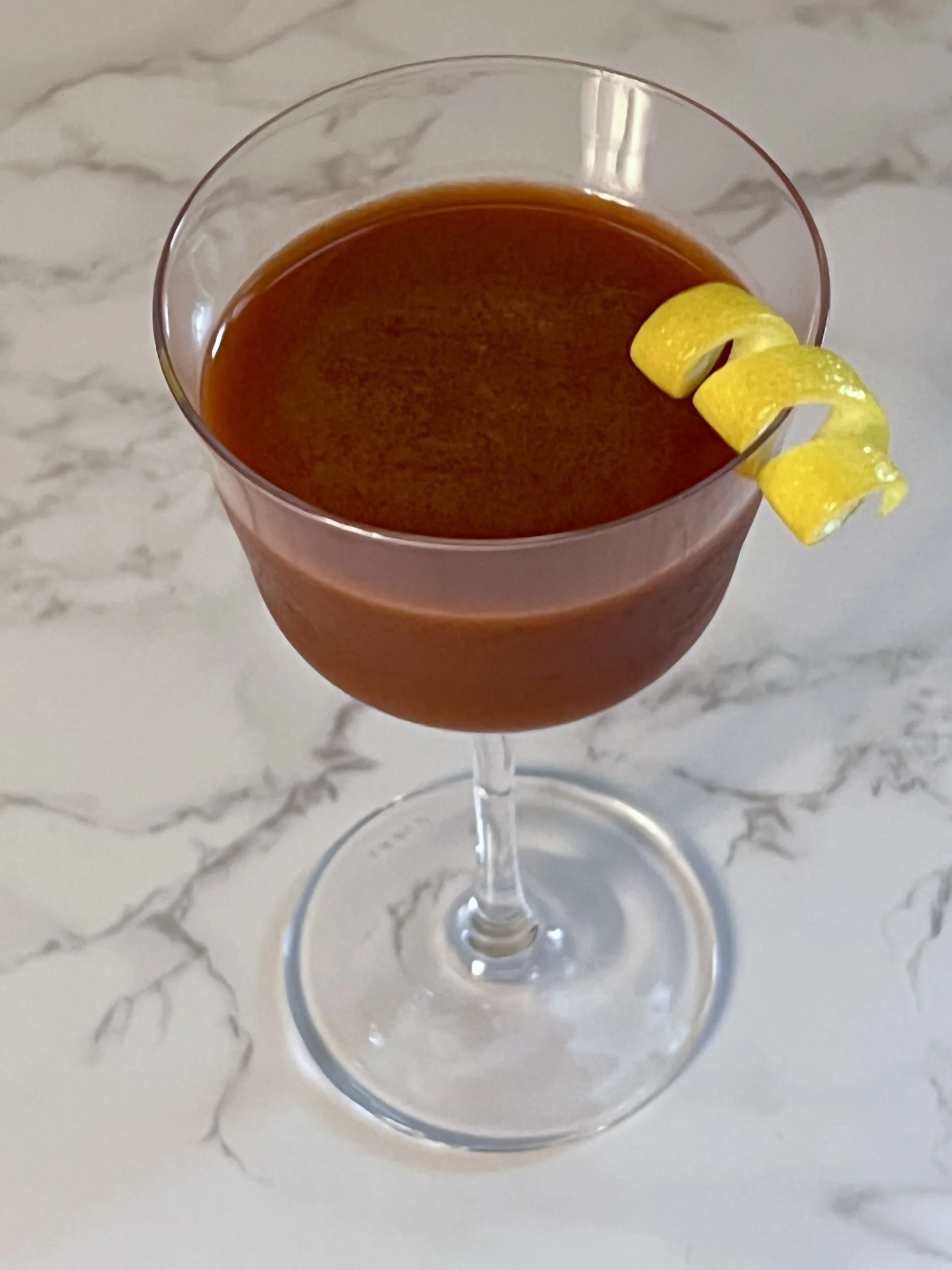 An up close angled aerial shot of a deep reddish-brown Trinidad Sour in a Nick & Nora glass garnished with a bright yellow lemon twist.