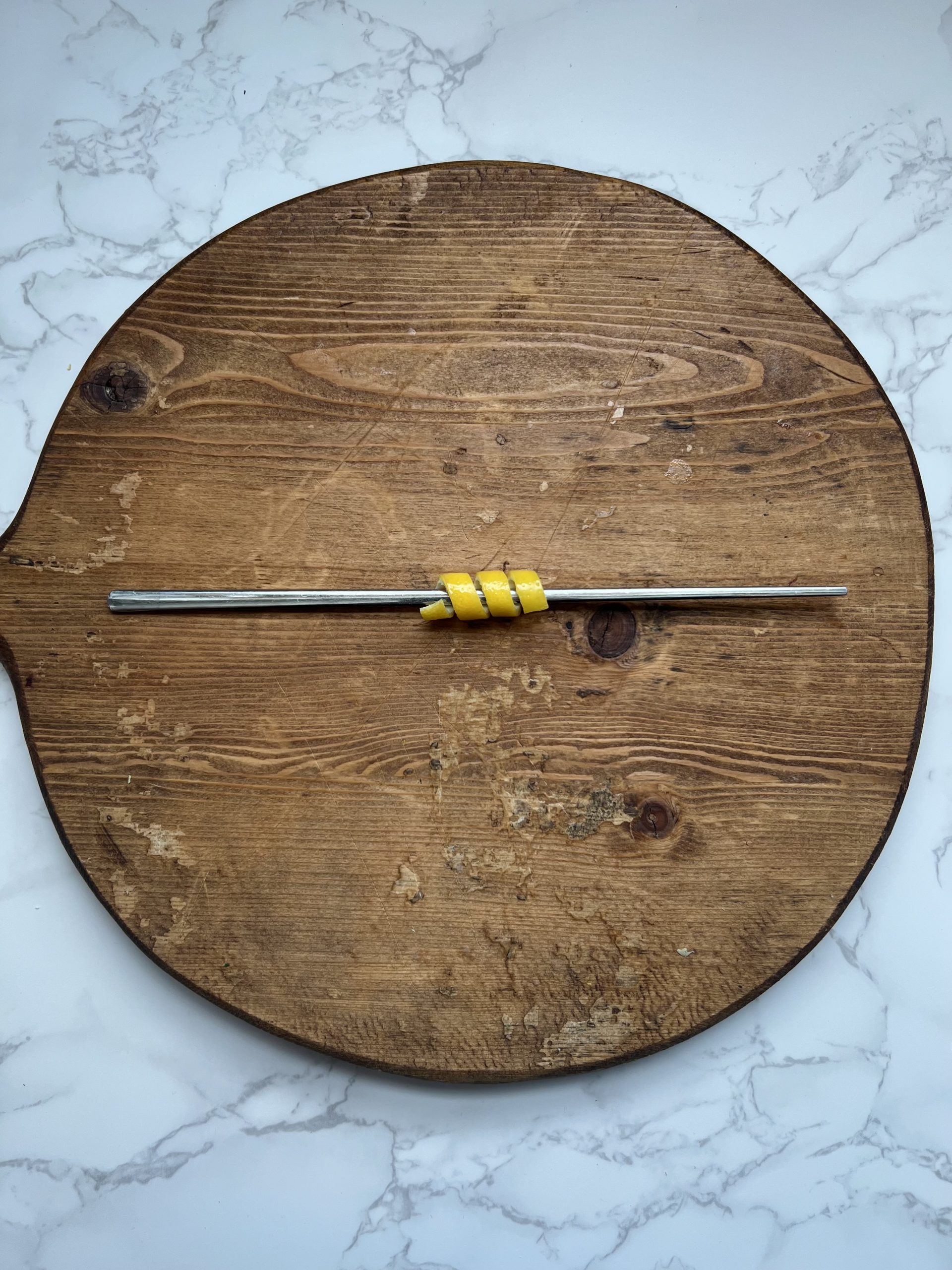 A lemon peel twisted around a metal chopstick on a cutting board.