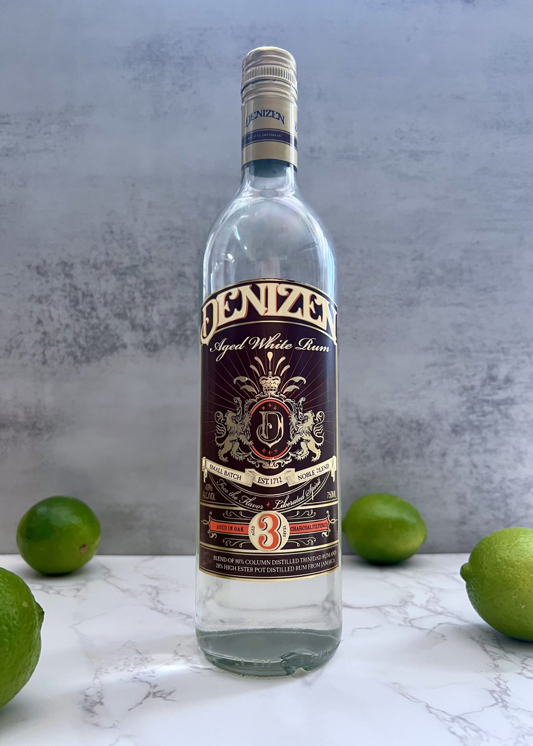 A bottle of Denizen 3 Year Aged White Rum on a counter surrounded by limes.