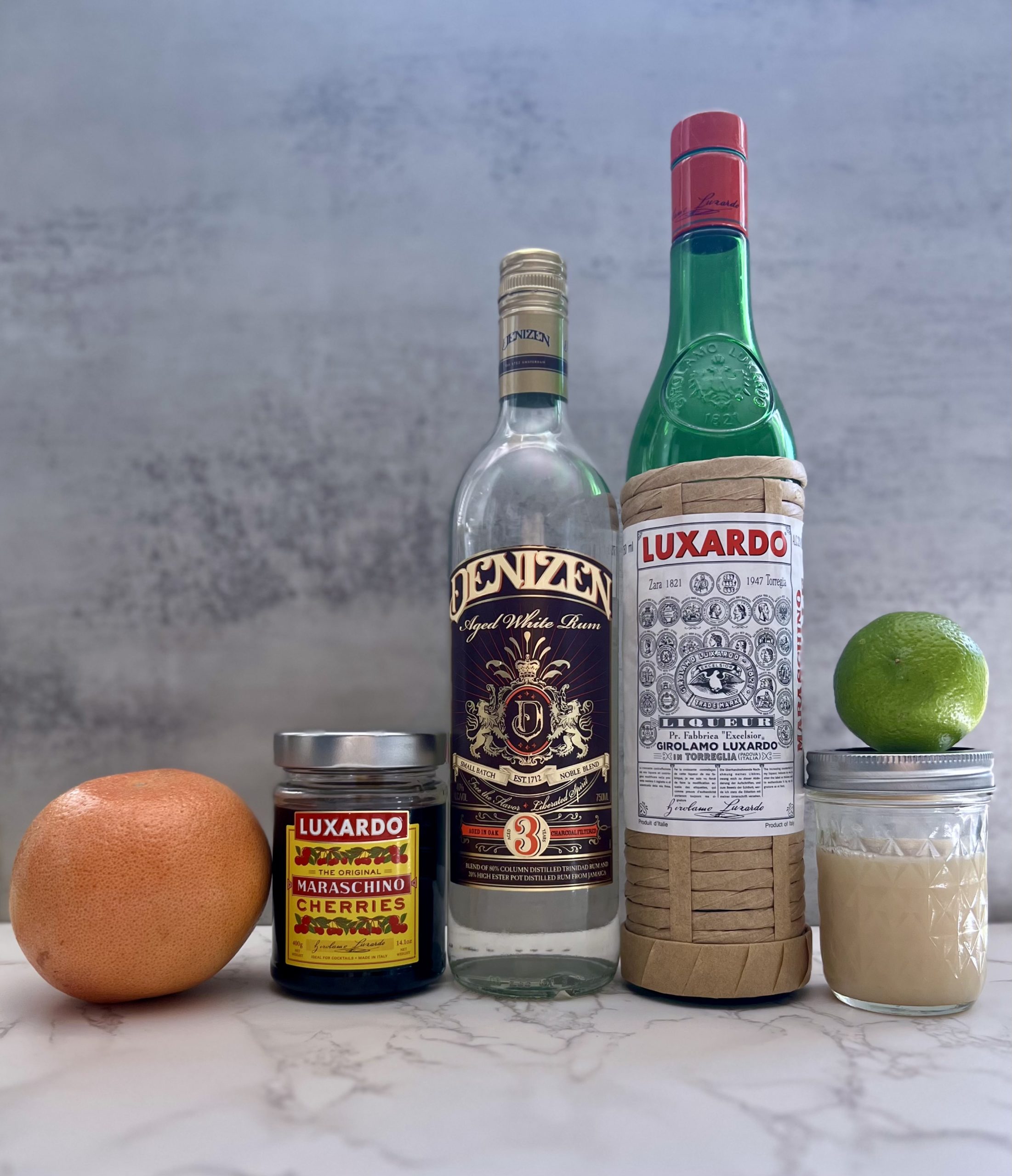 The ingredients for a Hemingway Daiquiri, including a grapefruit, Luxardo cherries, Denizen white rum, Luxardo Maraschino liqueur, orgeat almond syrup, and a lime. 