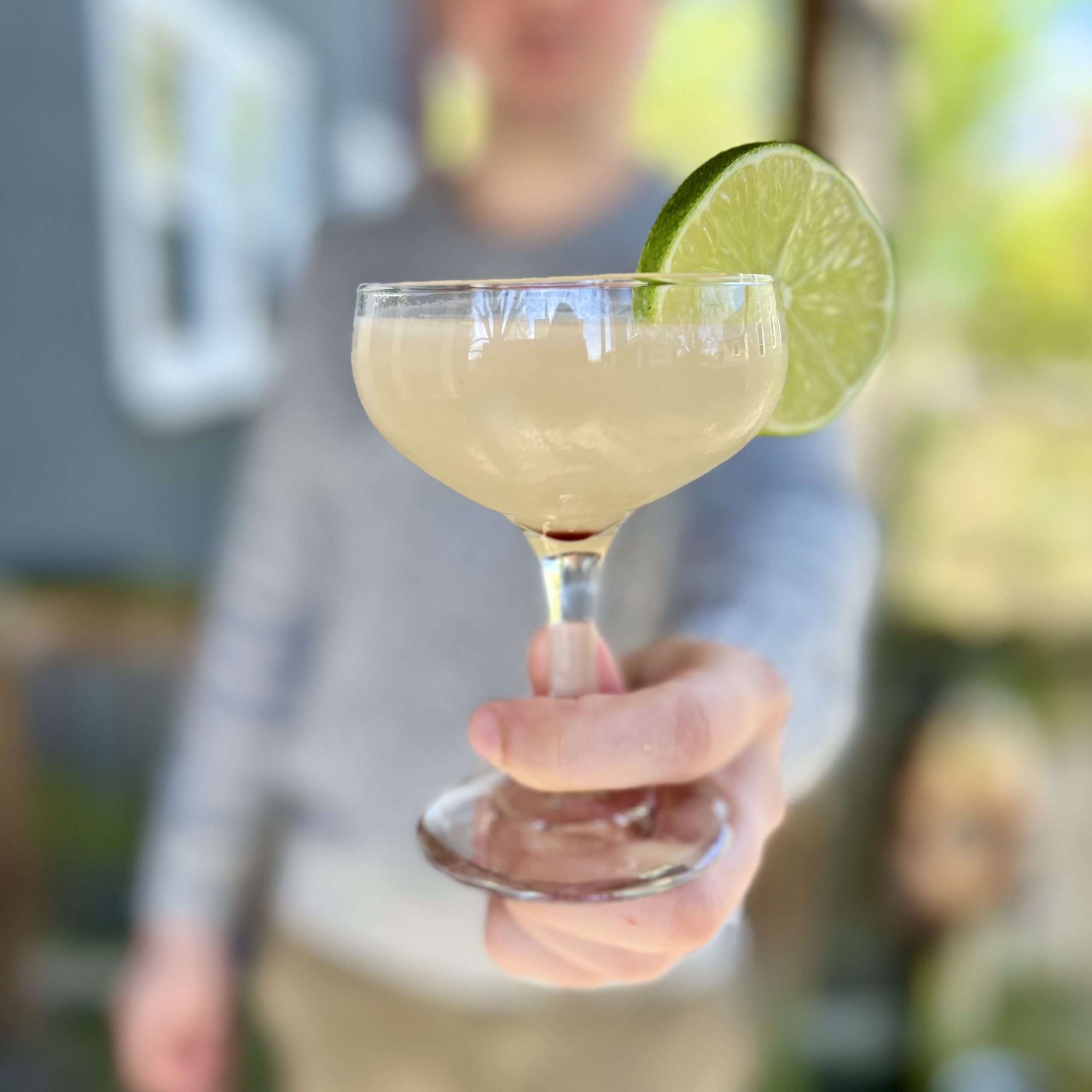 A Hemingway Daiquiri garnished with a lime held toward the camera held by a blurred out man in the background.
