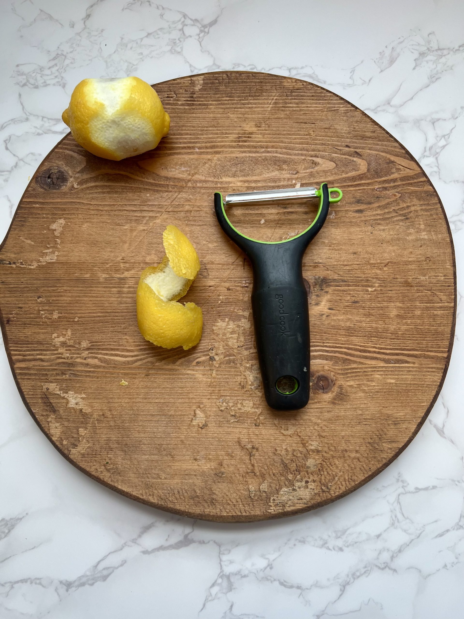 A vegetable peeler, large piece of lemon peel, and partially peeled lemon on a cutting board.