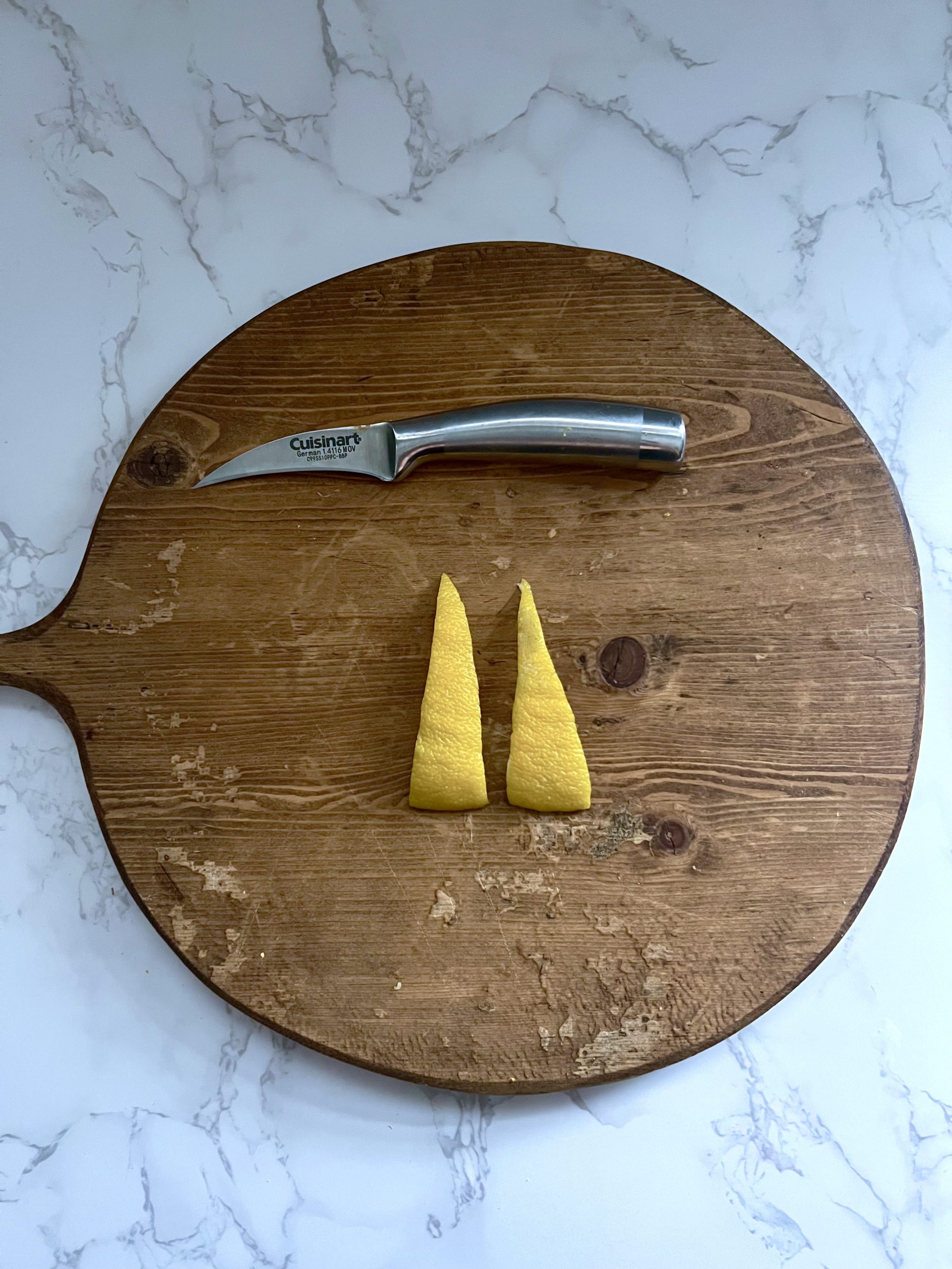 A knife with two long lemon peel triangles on a wooden cutting board.