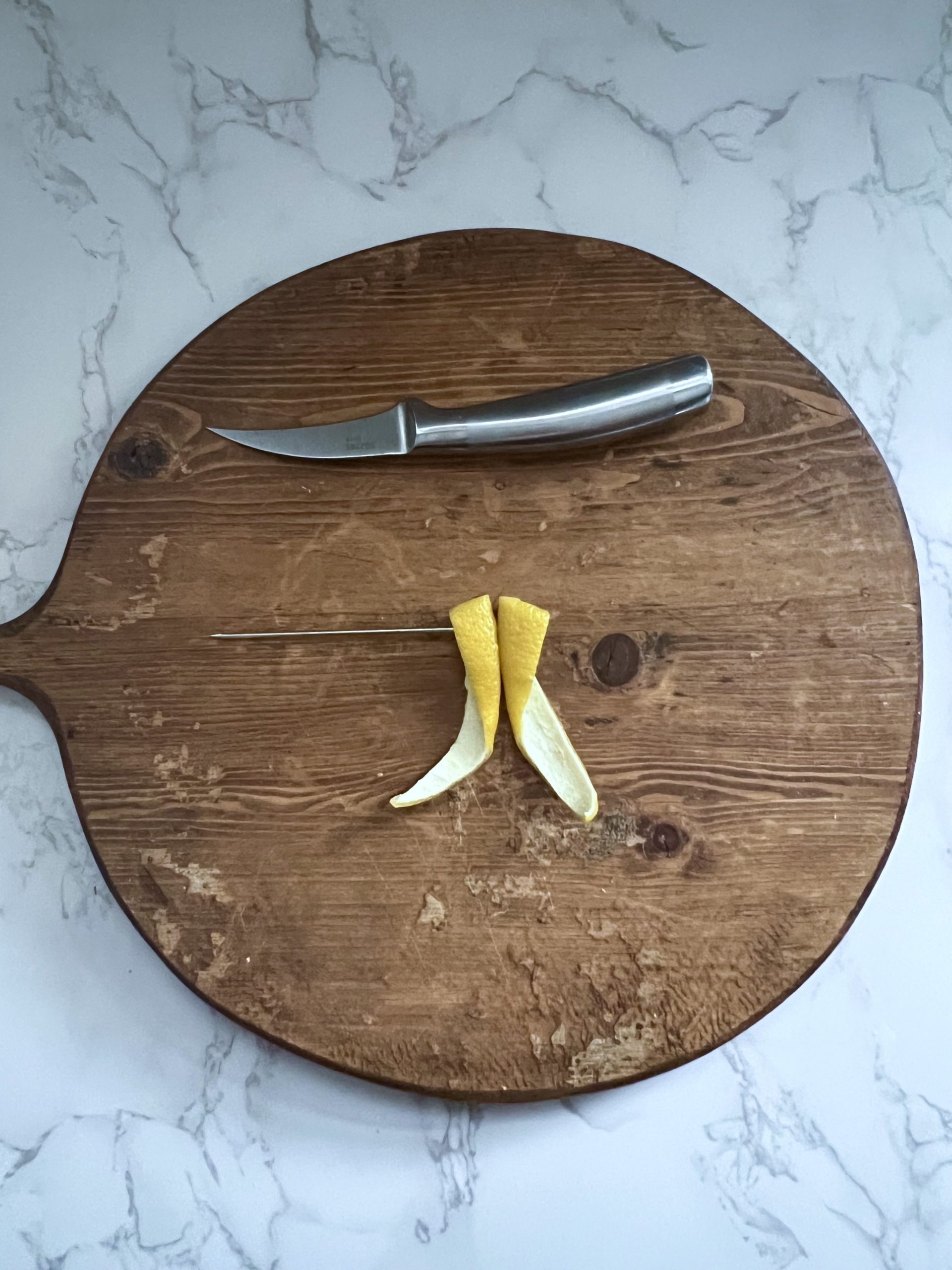 A knife and two pieces of lemon peel joined together with a cocktail pick to resemble the plane wings of the paper plane garnish on a cutting board.