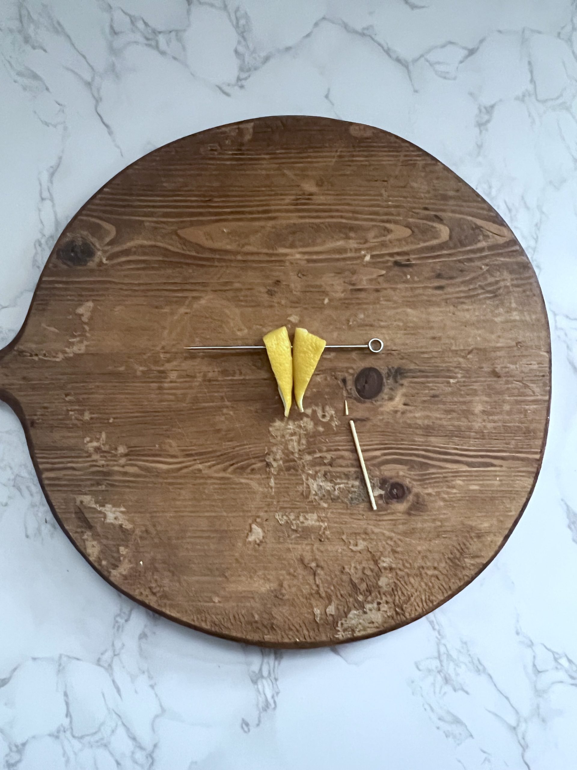 Two pieces of lemon peel joined together with a cocktail pick to resemble the plane wings of the paper plane garnish and a toothpick with the tip broken off on a cutting board.