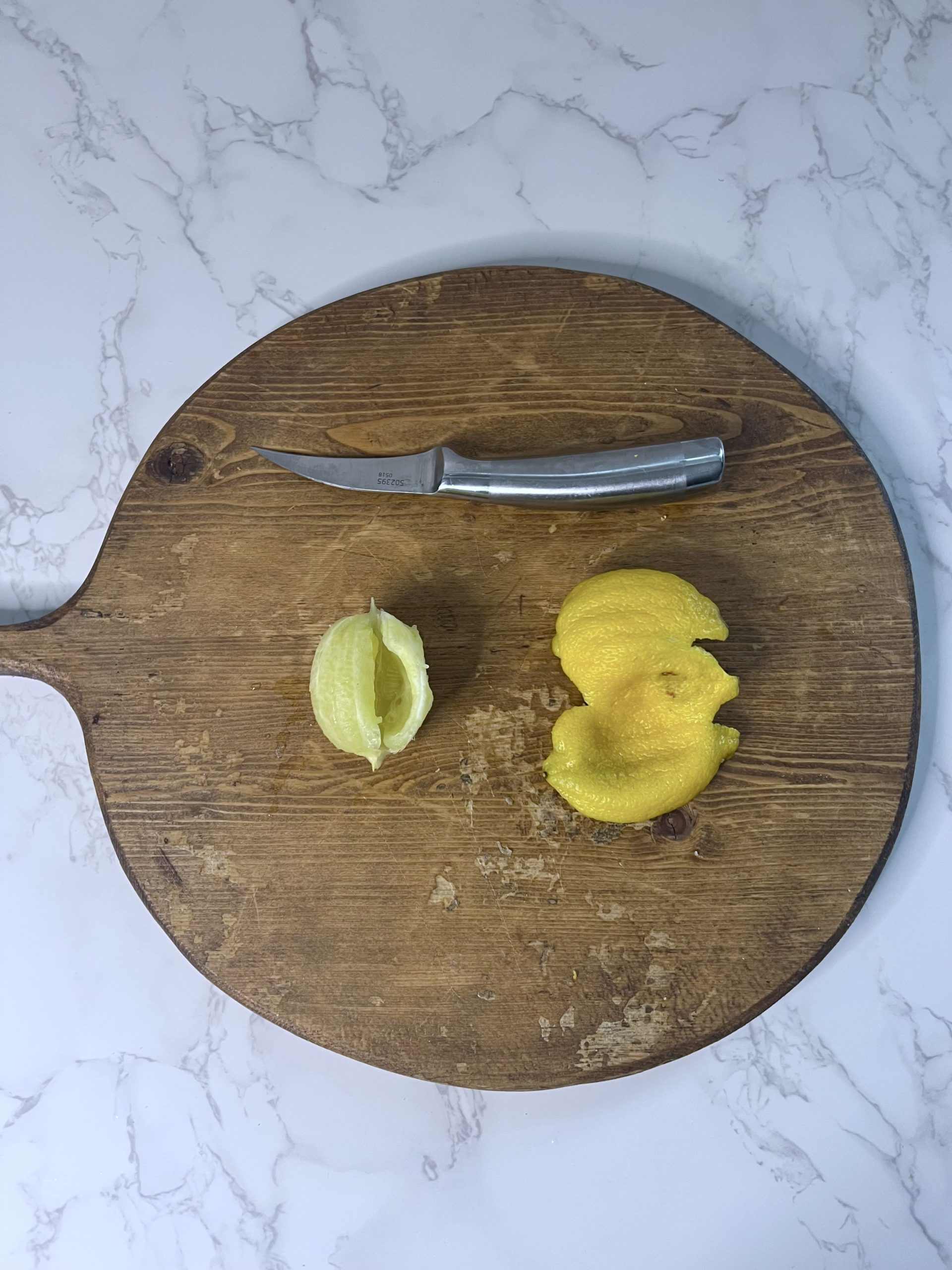 A knife, a peeled lemon, and an entire lemon peel on a wooden cutting board.