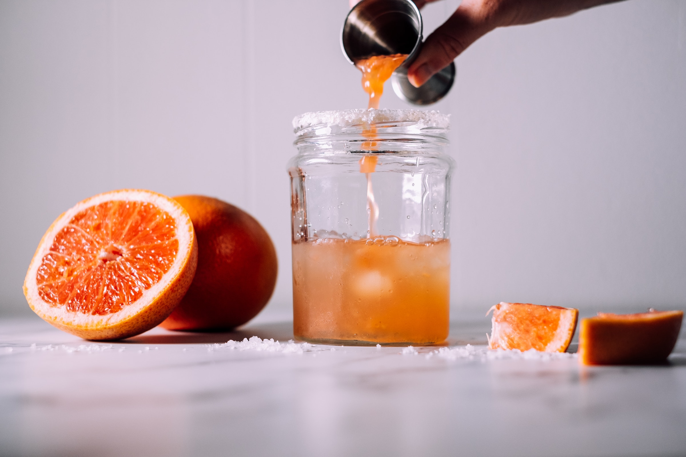 An image of someone pouring a jigger of grapefruit juice into a salt-rimmed glass to make a Salty Dog cocktail, surrounded by grapefruit slices